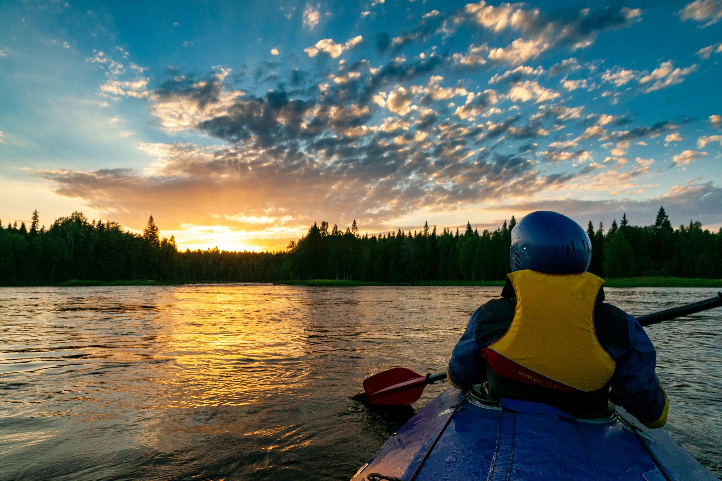 A person kayaking on a river at sunset, wearing a helmet and life jacket, with a forested shoreline and a sky filled with clouds illuminated by the setting sun.