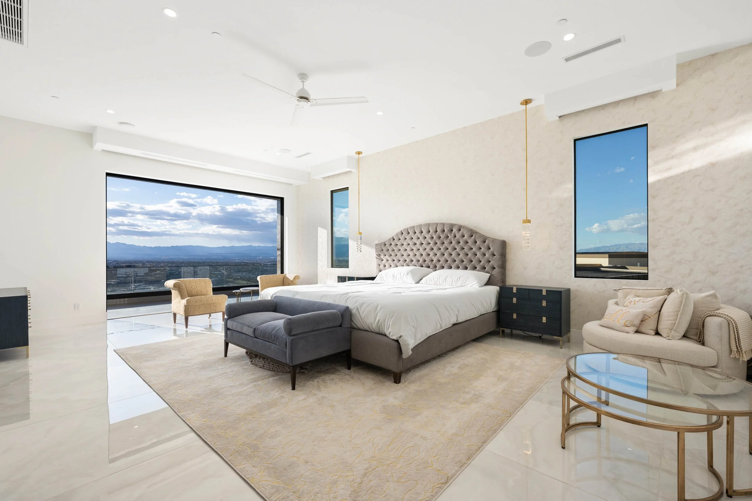 Modern bedroom with a large bed, tufted headboard, sitting area with chairs, and a view of mountains and sky through windows.