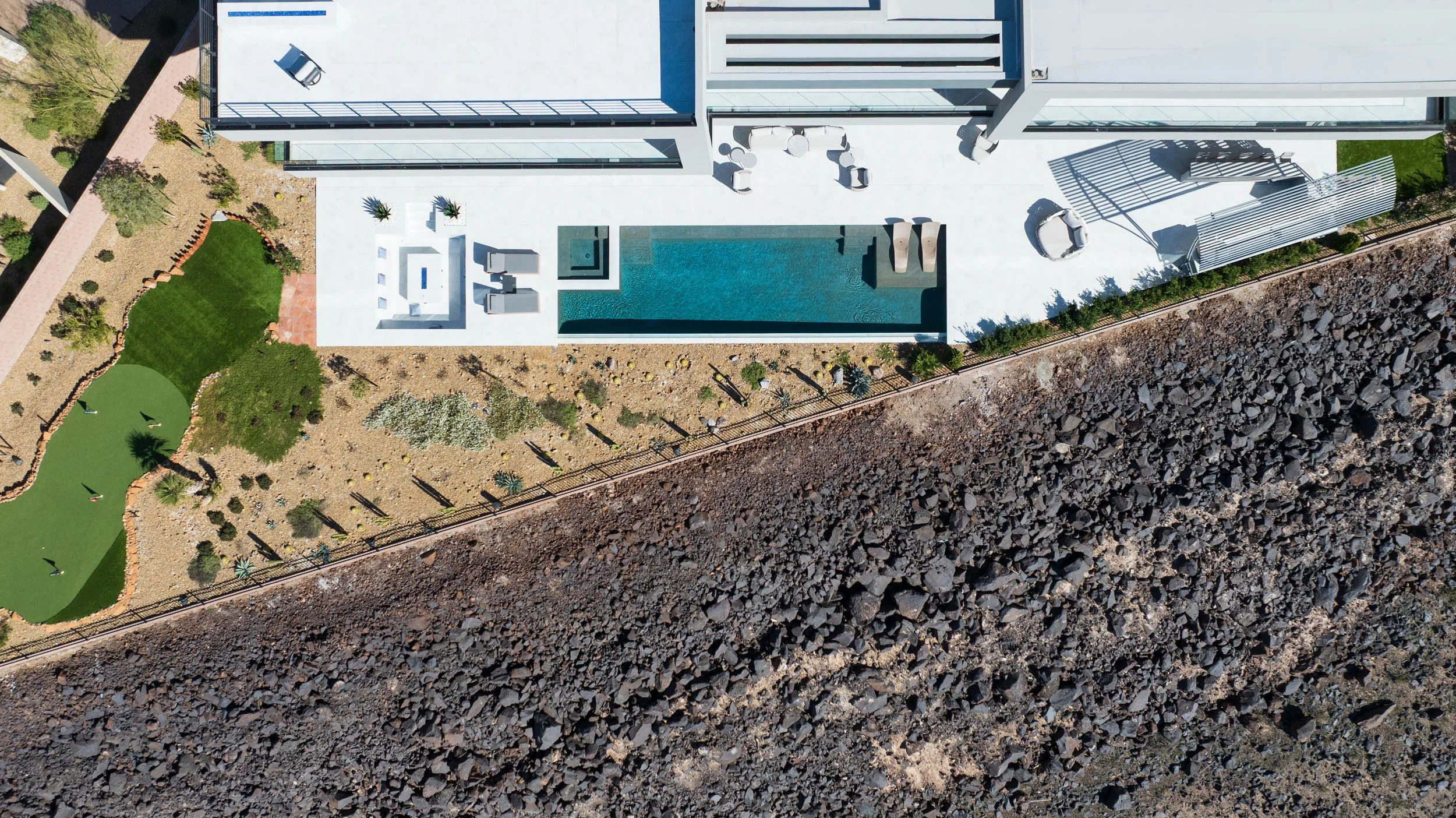 Aerial view of a modern house with a pool and outdoor seating area, next to a rocky terrain with sparse vegetation.