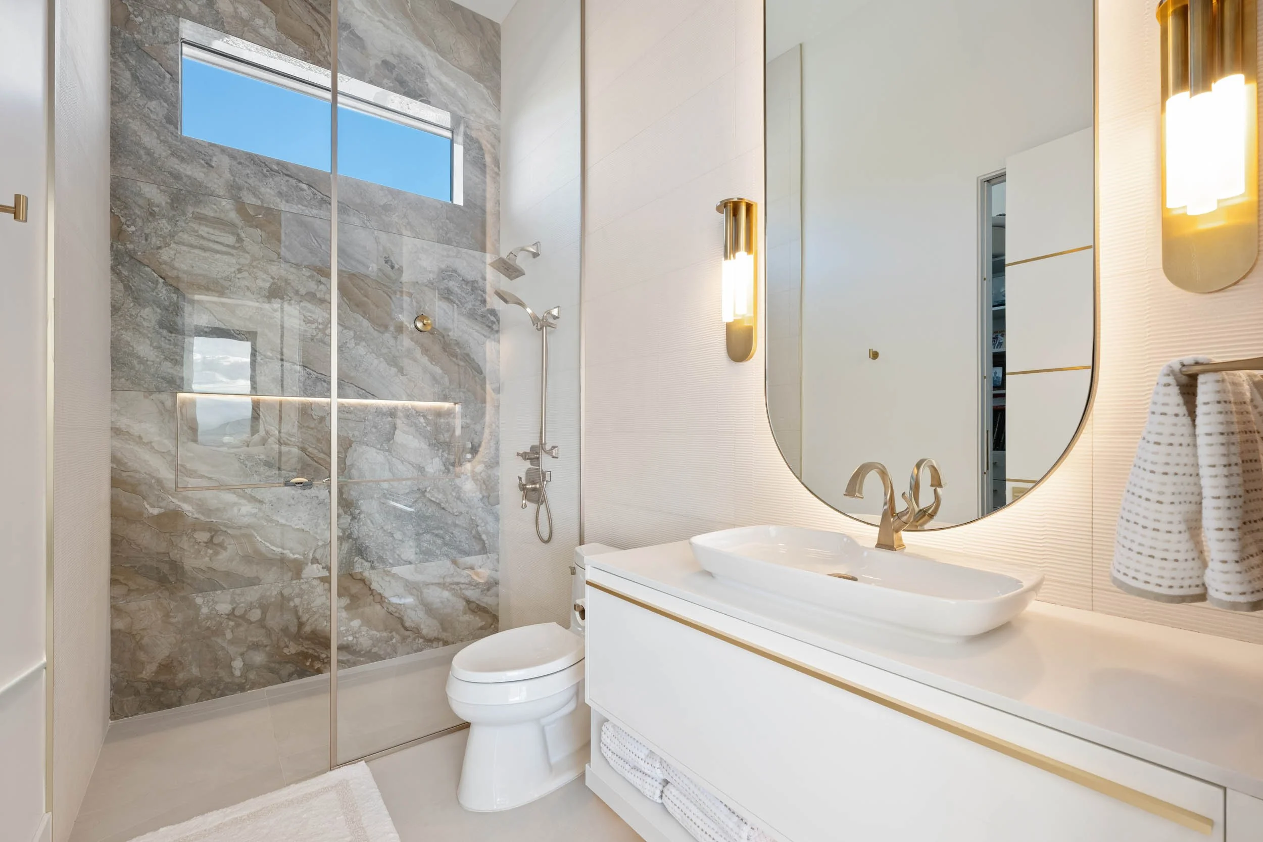 Modern bathroom with a walk-in shower featuring gray marble tiles, a small rectangular window, a white toilet, a white vanity with a round vessel sink, a large oval mirror, and gold wall-mounted light fixtures.