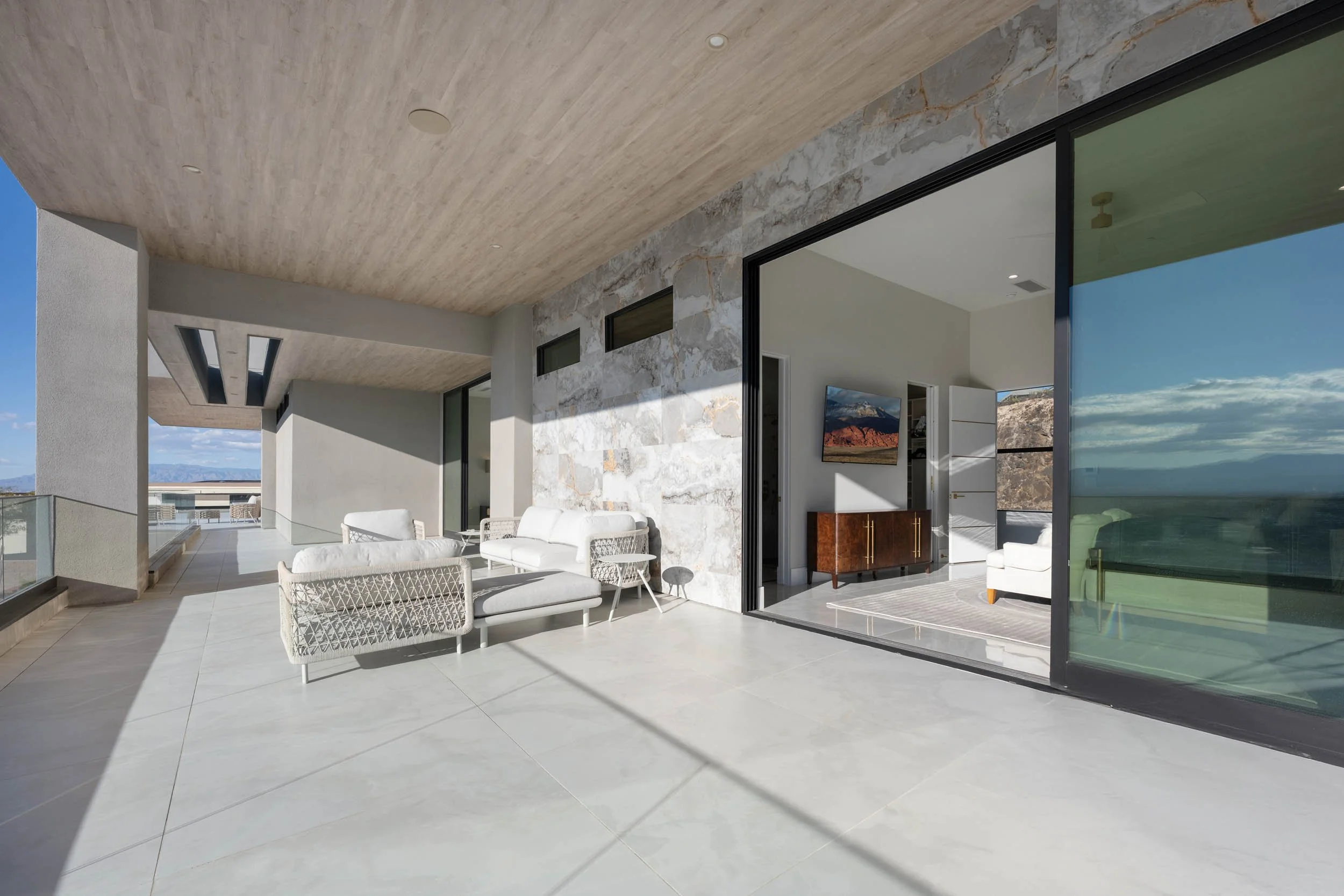Modern outdoor balcony with white seating and glass railing overlooking mountains and sky.