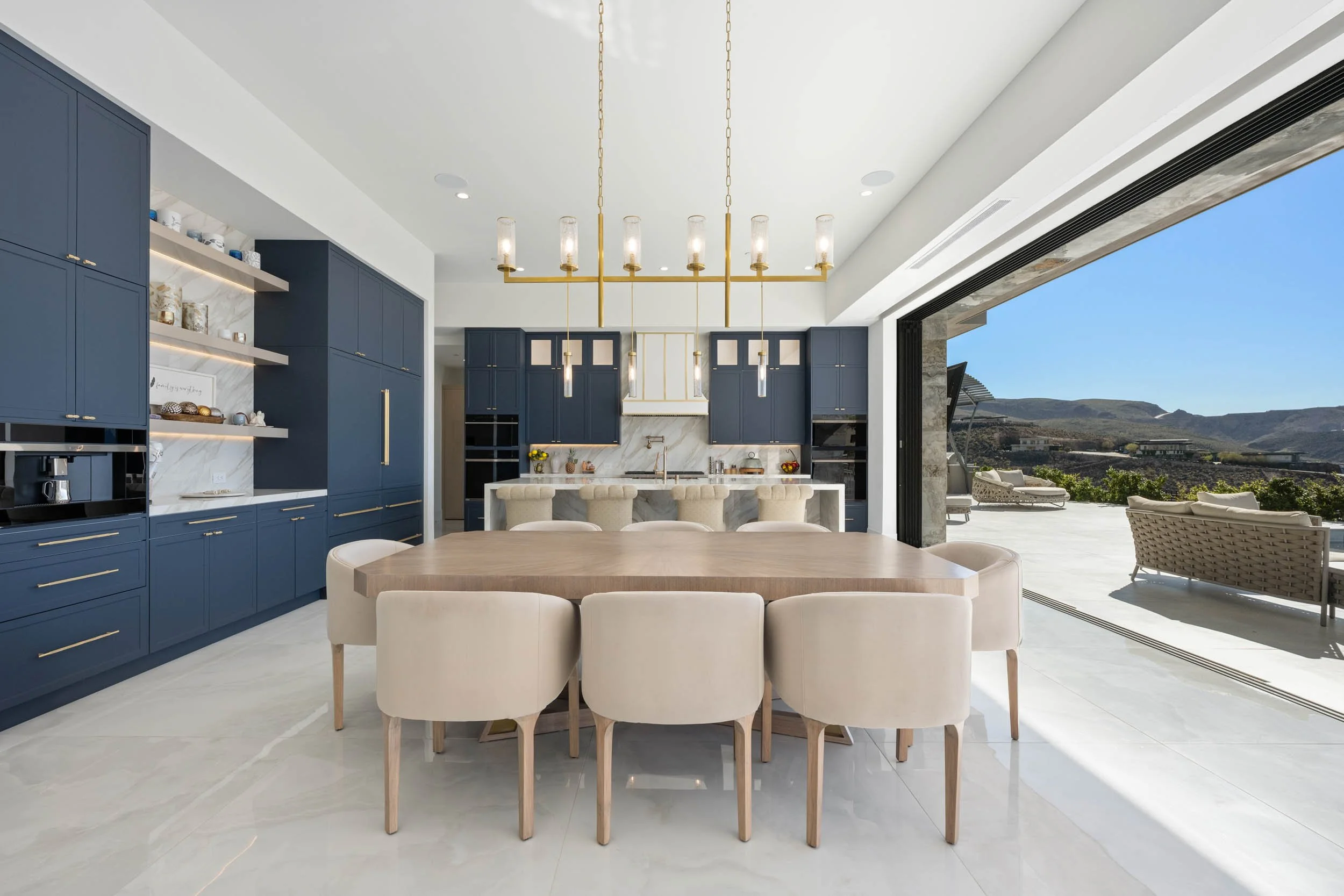 Modern kitchen with blue cabinets, white marble backsplash, large marble island with bar stools, beige dining chairs around a wooden table, large open sliding door revealing outdoor patio and mountain view.