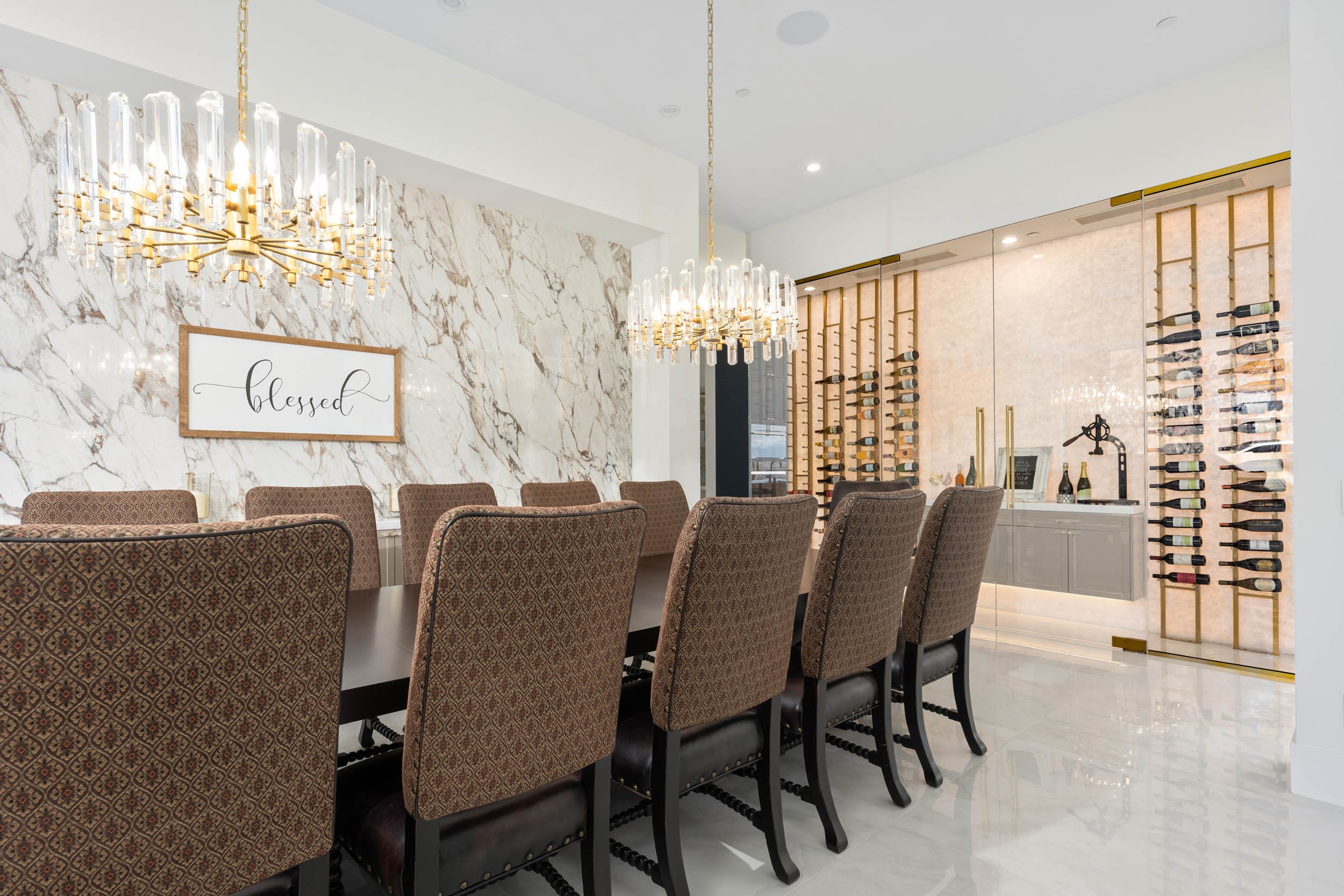Elegant dining room with a long table surrounded by upholstered chairs, a marble accent wall with a framed sign that says 'blessed,' two large chandeliers, and a wall wine rack with bottles displayed.