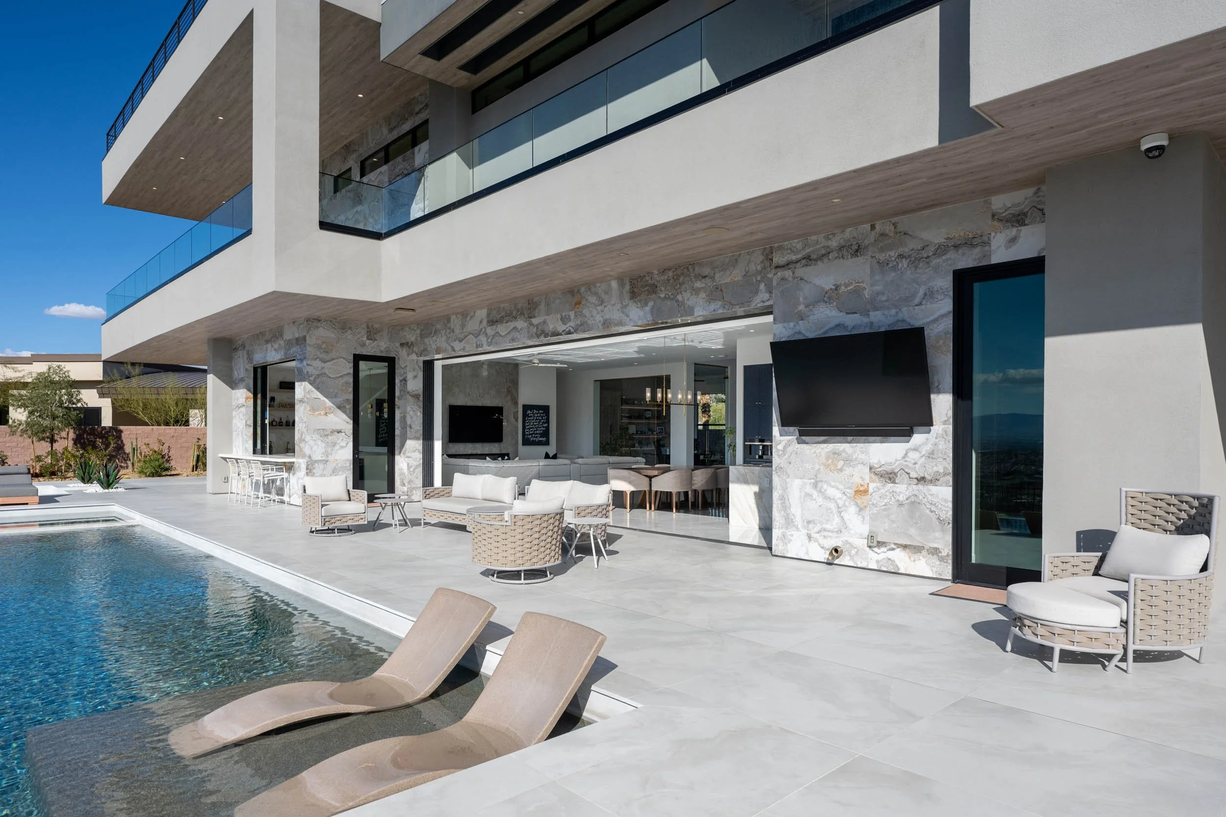 Modern residential outdoor patio with pool, lounge chairs, and outdoor seating area under a large building with balconies, glass railings, and stone accents.