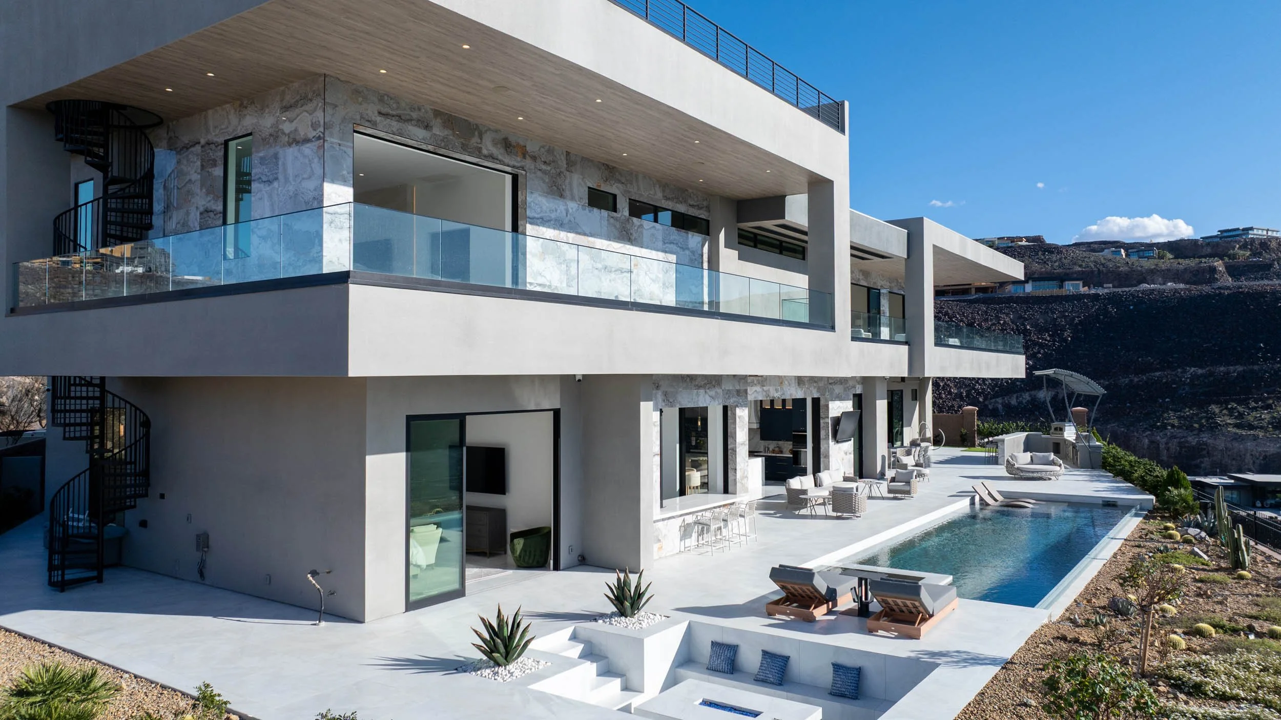 Modern multi-story house with a swimming pool on a sunny day, featuring glass railings, outdoor seating, and desert landscape surroundings.