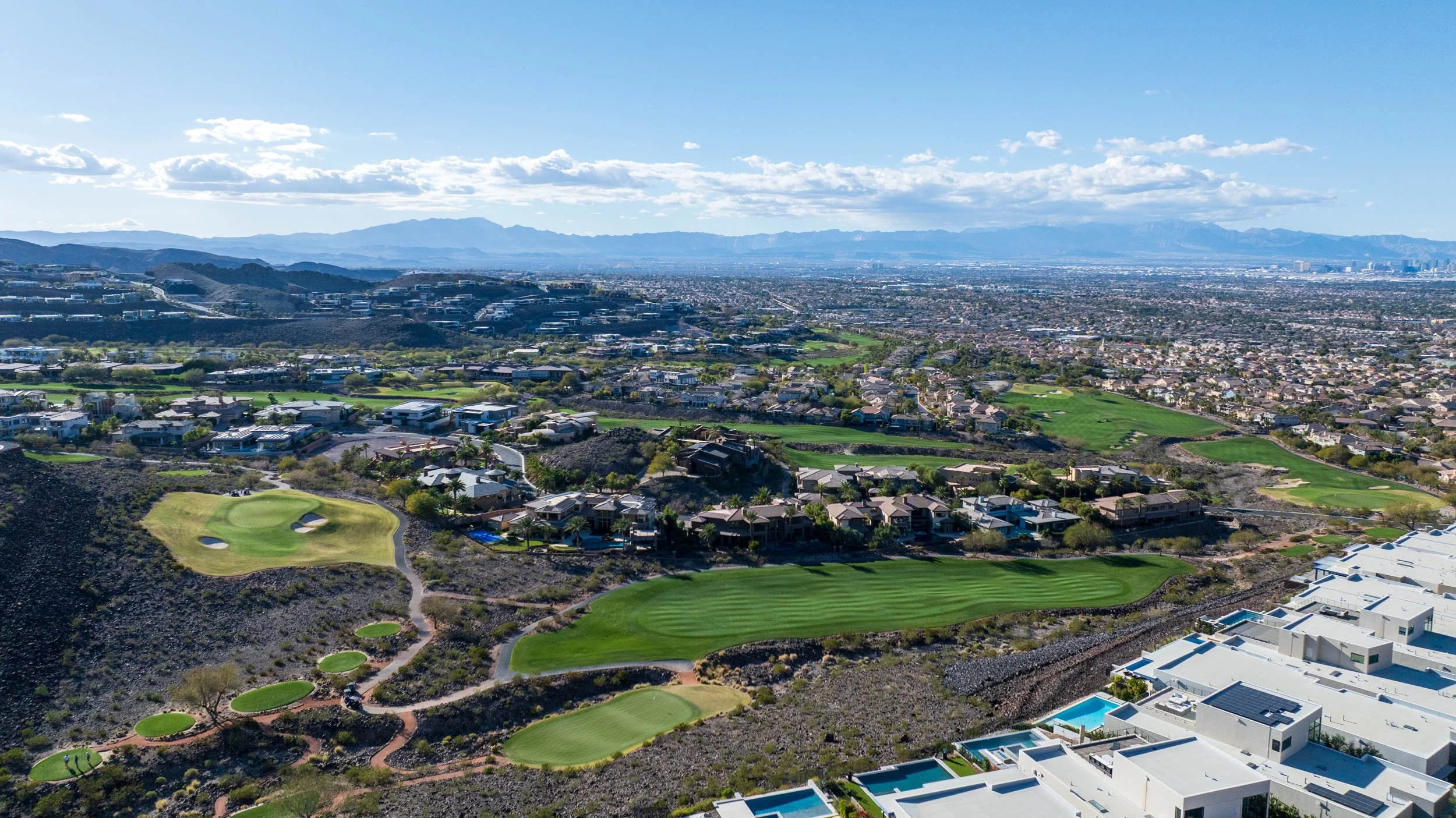 Aerial view of a golf course surrounded by houses, with a cityscape and mountains in the distance under a partly cloudy sky.