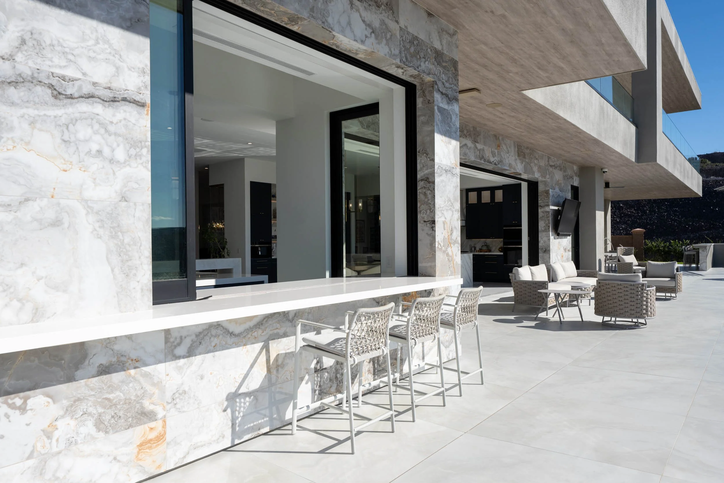 Modern outdoor patio with lounge chairs and sofa, adjacent to a house with large windows and marble exterior, overlooking a mountain landscape.
