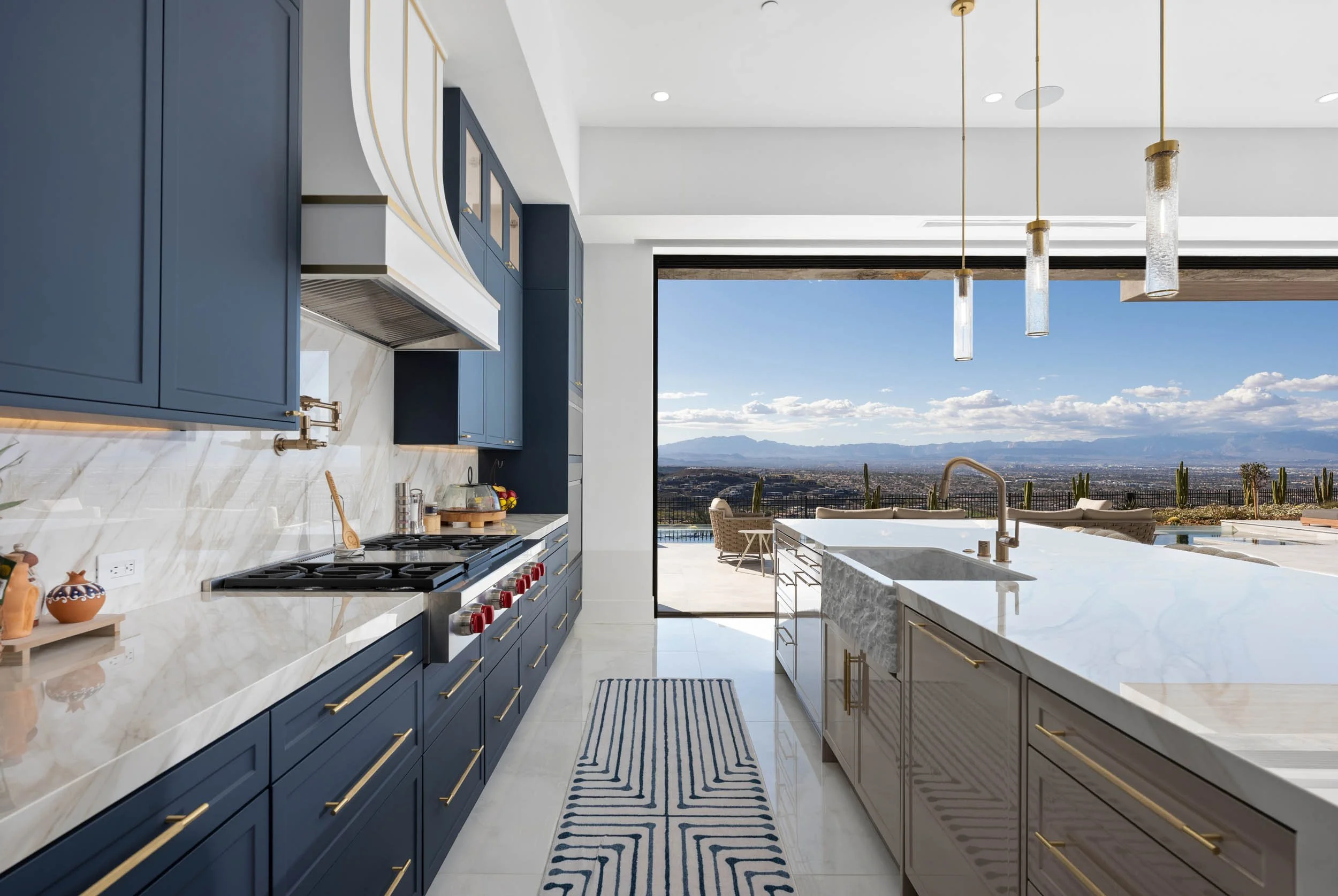 Modern kitchen with navy blue and beige cabinets, white marble countertops, and a view of a desert landscape with mountains and cacti through large open sliding doors. Hanging pendant lights above the island, a striped rug on the floor.