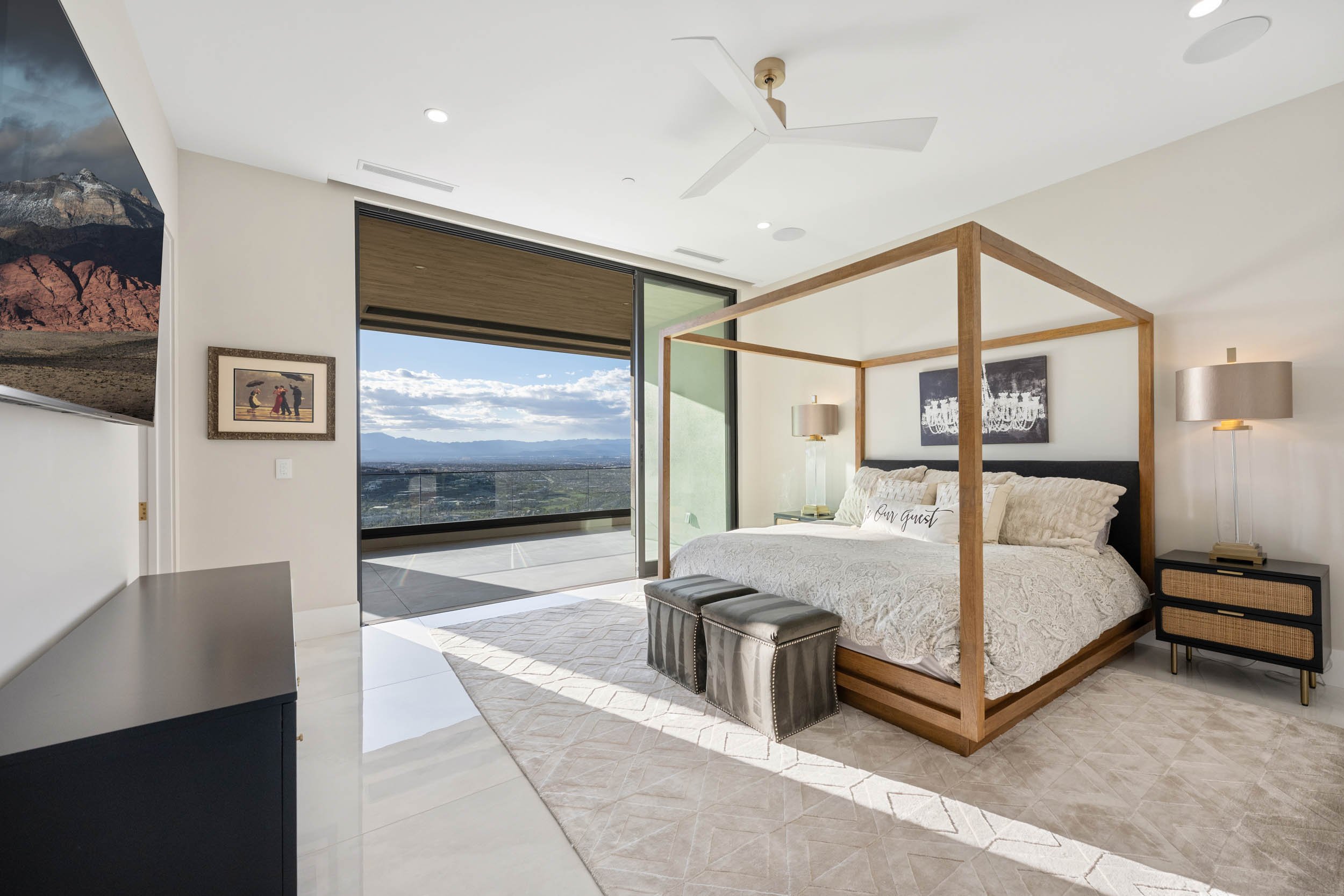 Modern bedroom with a four-poster bed, nightstands with lamps, a piece of wall art, an ottoman, and a sliding glass door leading to a balcony with a city view.