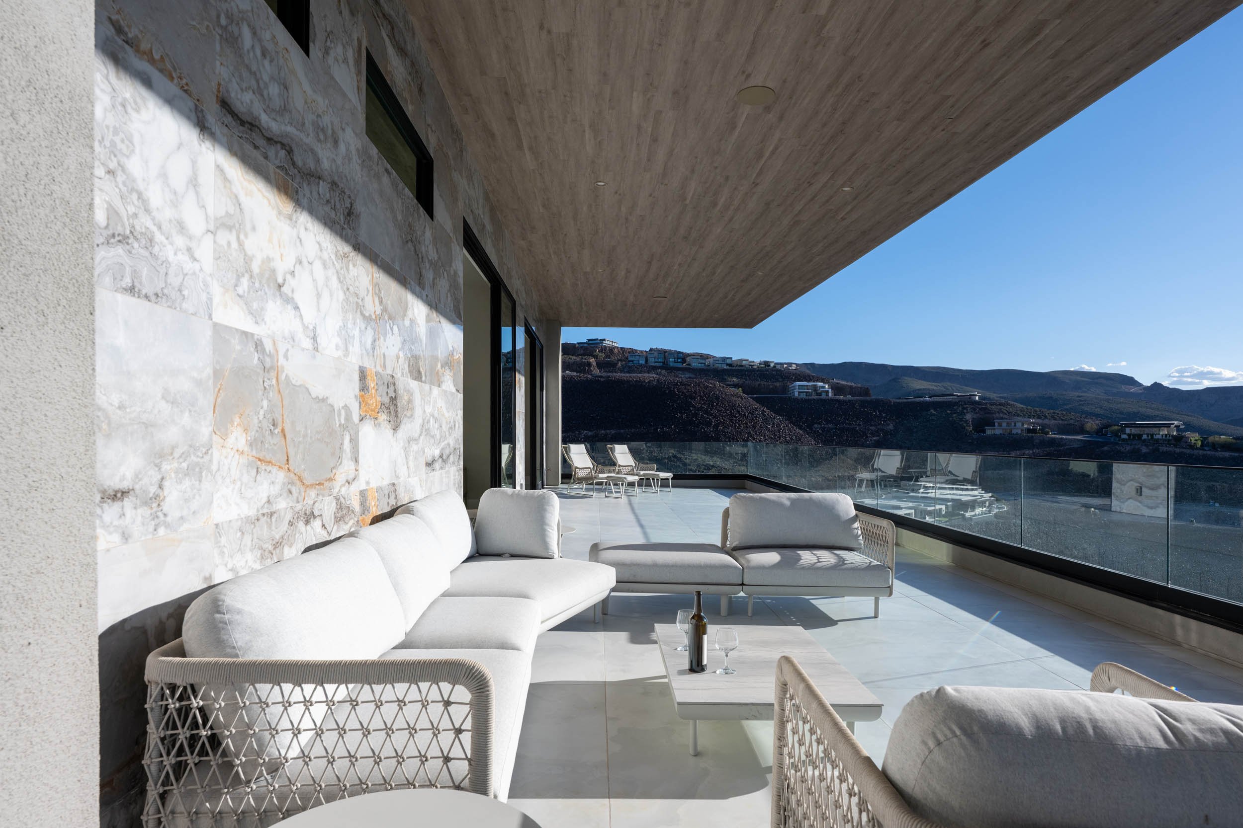 Contemporary outdoor balcony with white sectional sofas, wicker armchairs, and a coffee table with a bottle and glasses, overlooking a hilly landscape and a clear blue sky.