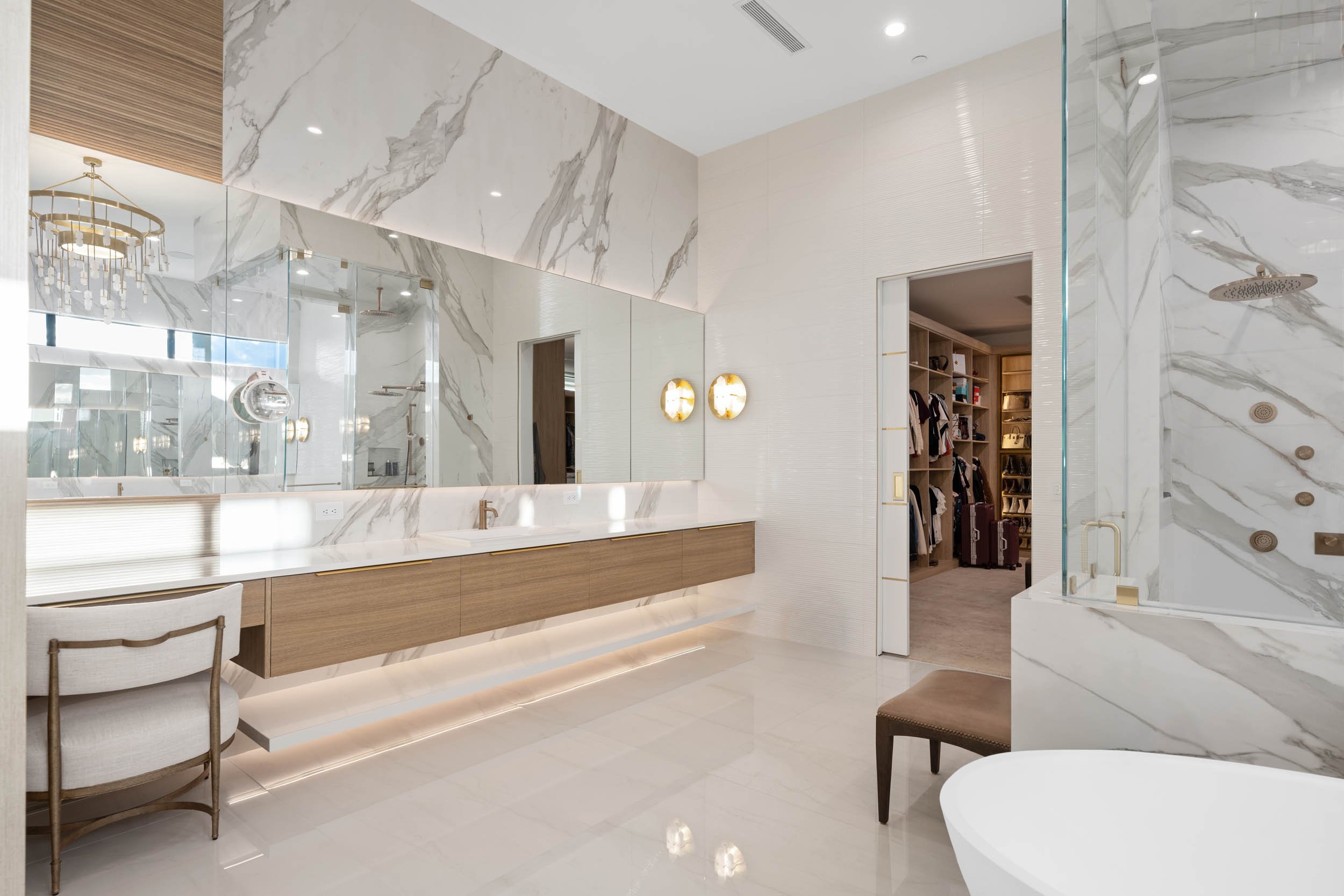 Luxurious bathroom with marble walls and floors, a large mirror above a long wooden vanity with a white countertop, modern wall sconces, and a walk-in closet visible through a doorway.