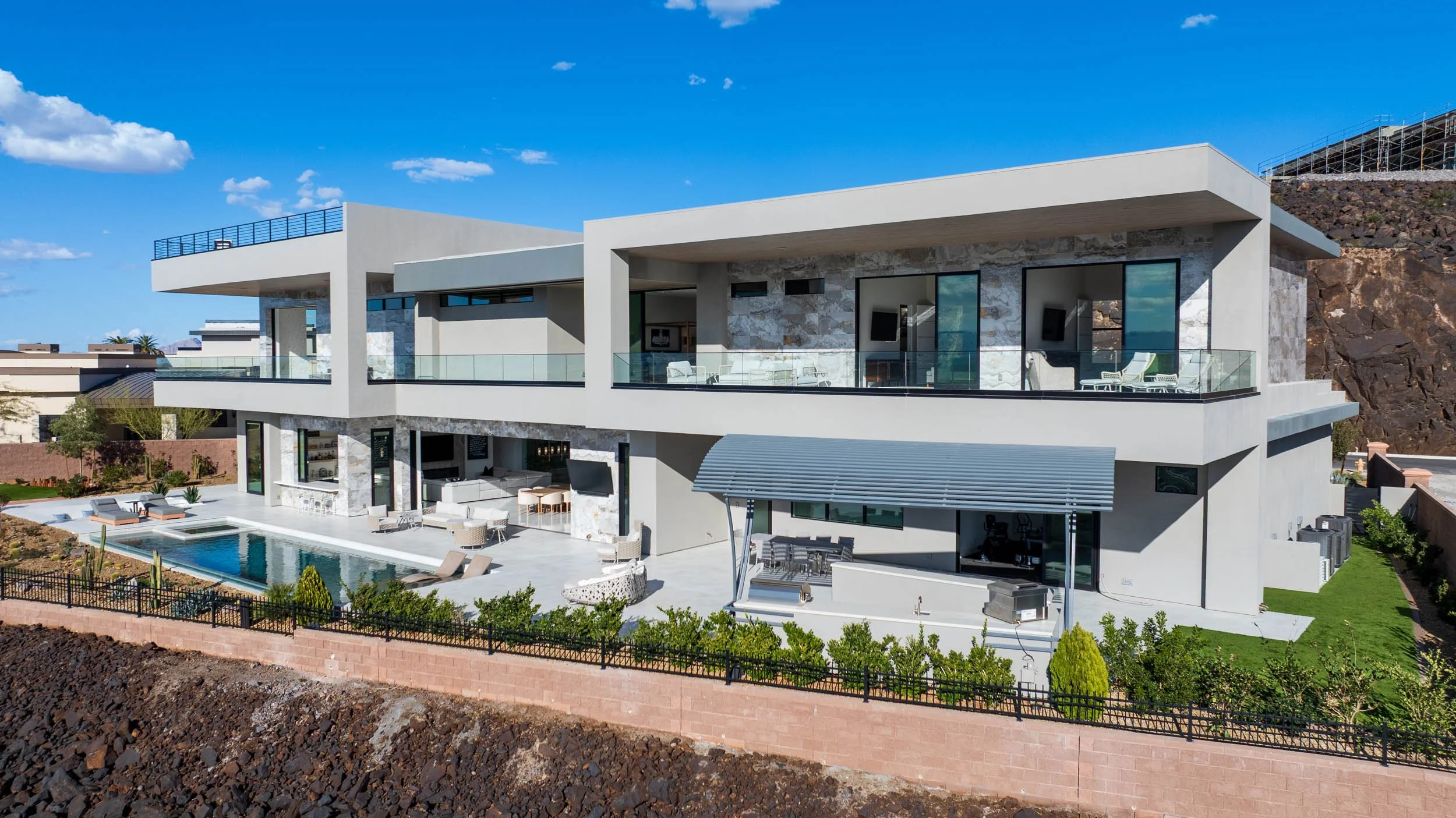 Modern multi-story house with large glass windows, balconies, poolside patio with lounge chairs, and landscaped yard, set against a rocky hillside and clear blue sky.