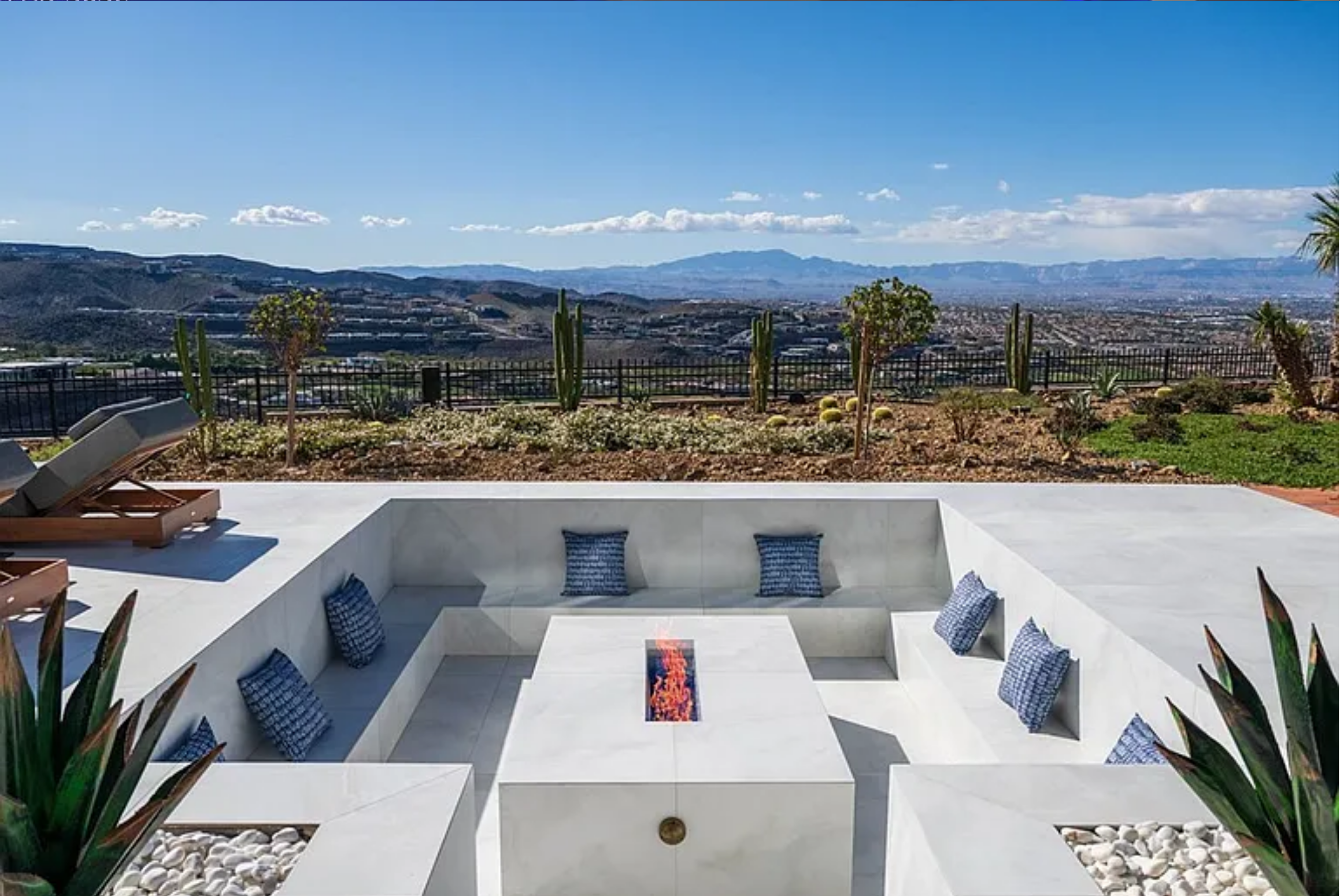 Outdoor seating area with built-in benches and blue patterned cushions surrounds a rectangular fire pit, with desert plants like cacti and succulents in the background against a mountain and cityscape view under a clear blue sky.
