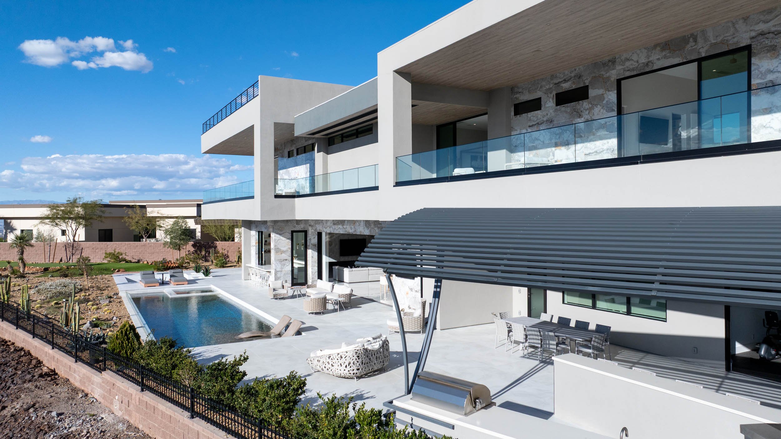 Modern house with a swimming pool, outdoor seating, and a shaded patio, under a blue sky with scattered clouds.