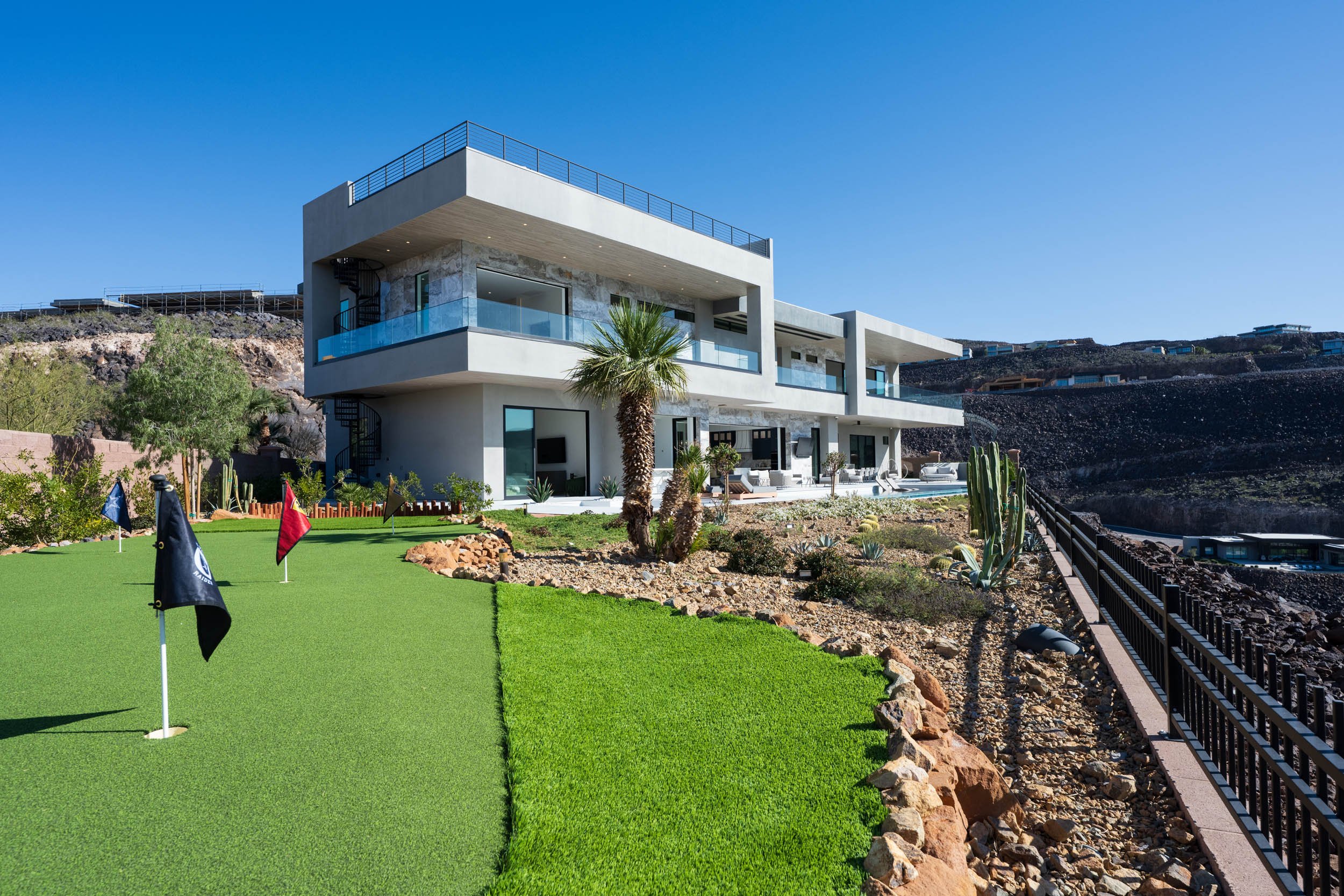 Modern multi-story house with large glass windows and balconies, landscaped front yard with artificial putting green, palm trees, cacti, and desert plants, and a metal fence along rocky terrain, under a clear blue sky.