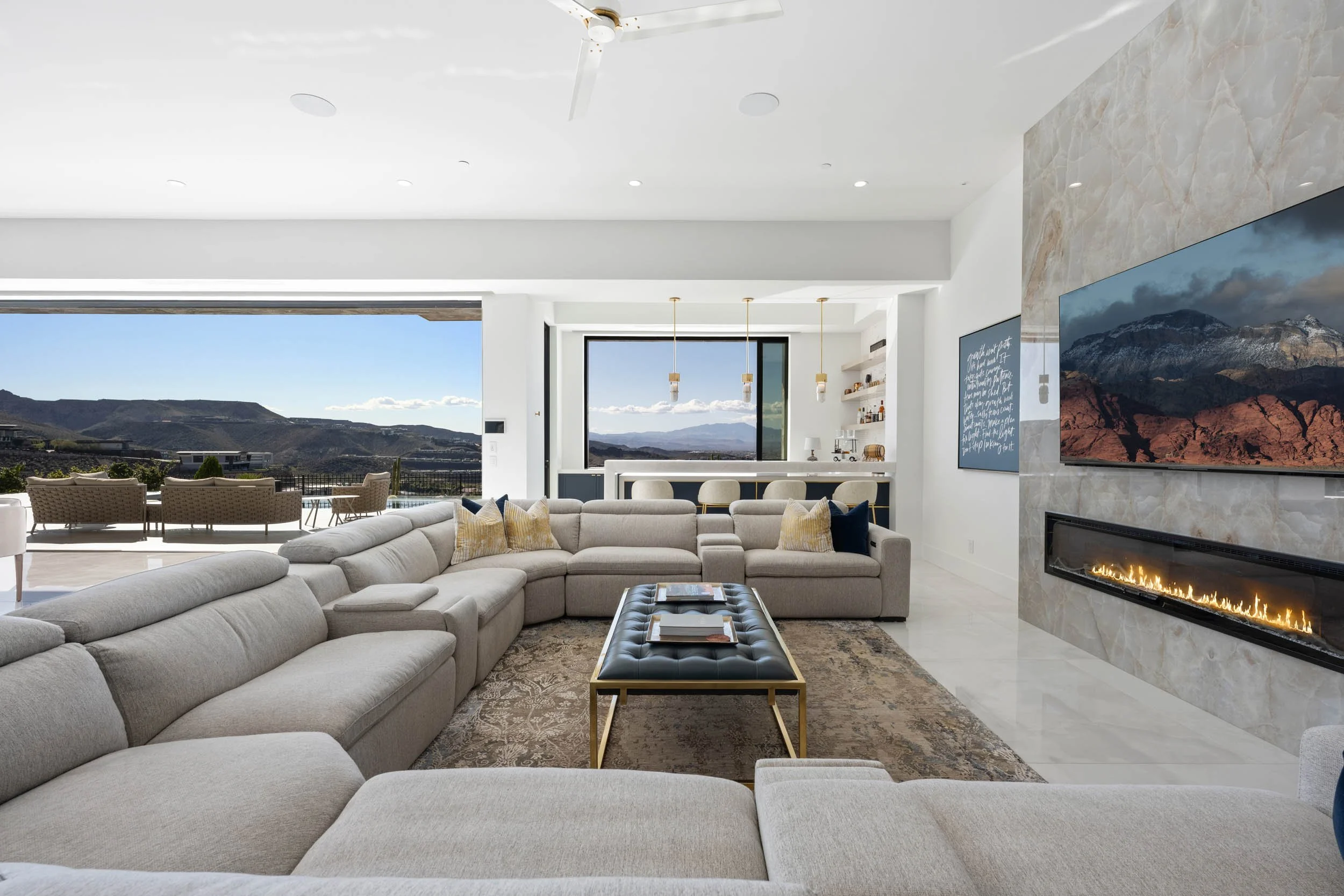 Modern living room with large beige sectional sofa, black leather coffee table, fireplace, mounted flat-screen TV, and large windows showing mountain views.