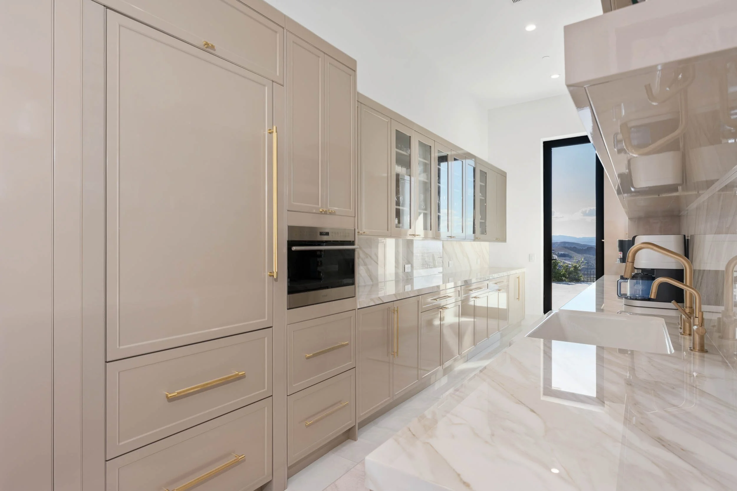 Modern kitchen with beige cabinets with gold handles, marble countertops, and a view outside through a glass door.