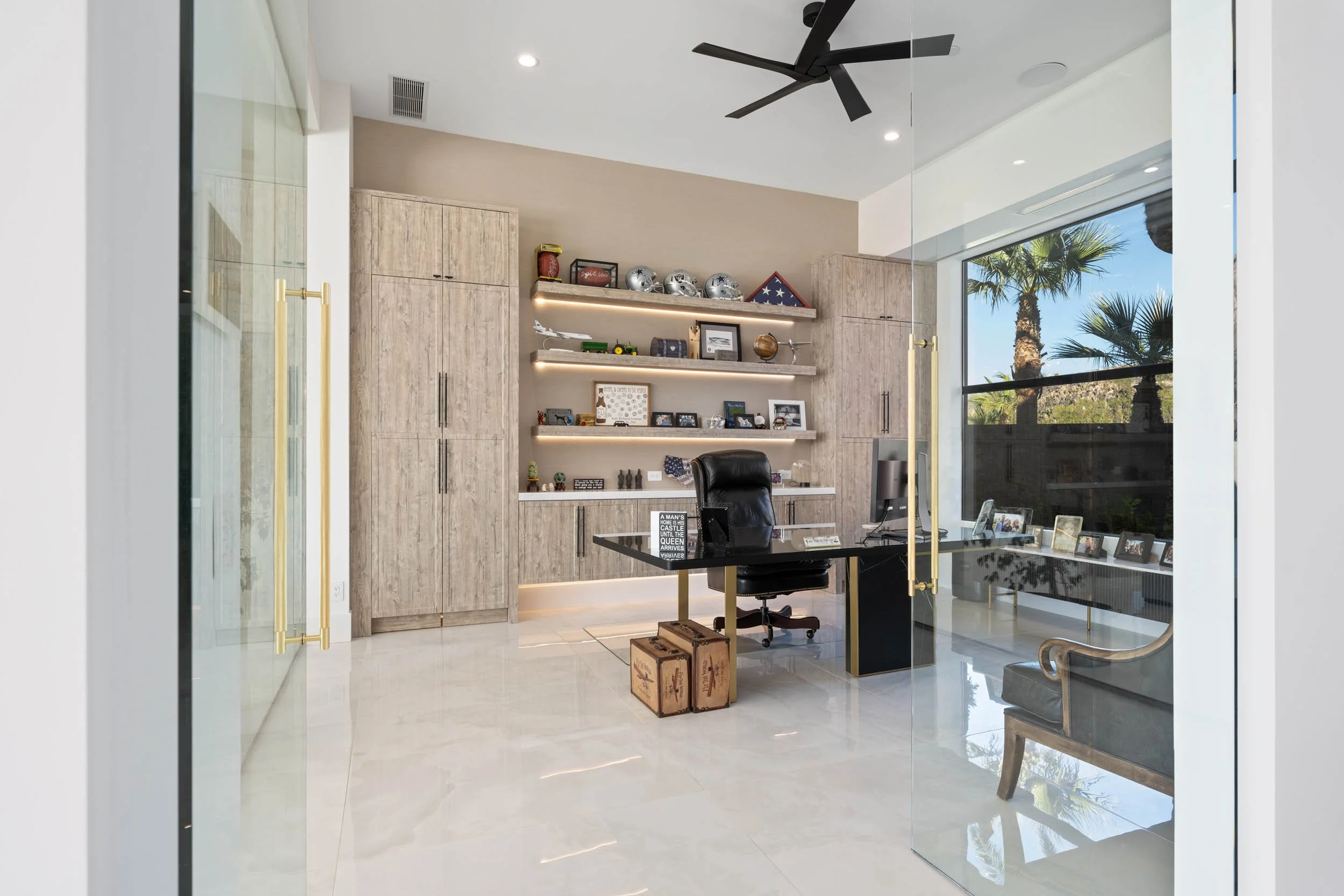 Modern office with glass doors, a black desk, a black office chair, wooden cabinets, a gray armchair, and shelves with various memorabilia, photos, and decorative items.