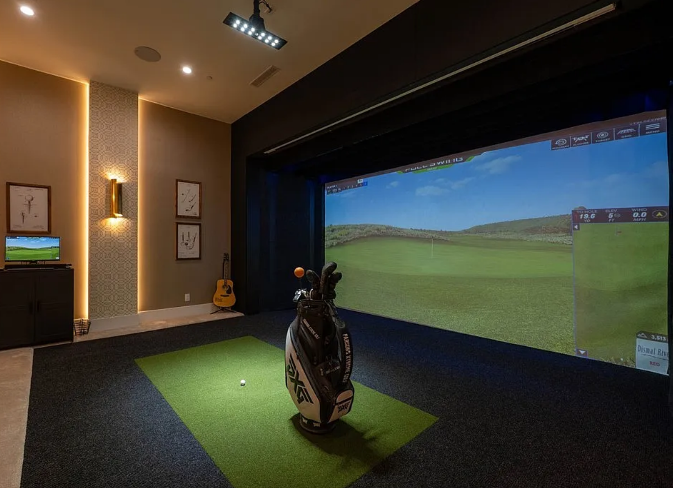 Indoor golf simulator room with a large screen displaying a golf course, a golf bag with clubs and a golf ball on a green mat, and a guitar against the wall to the left.