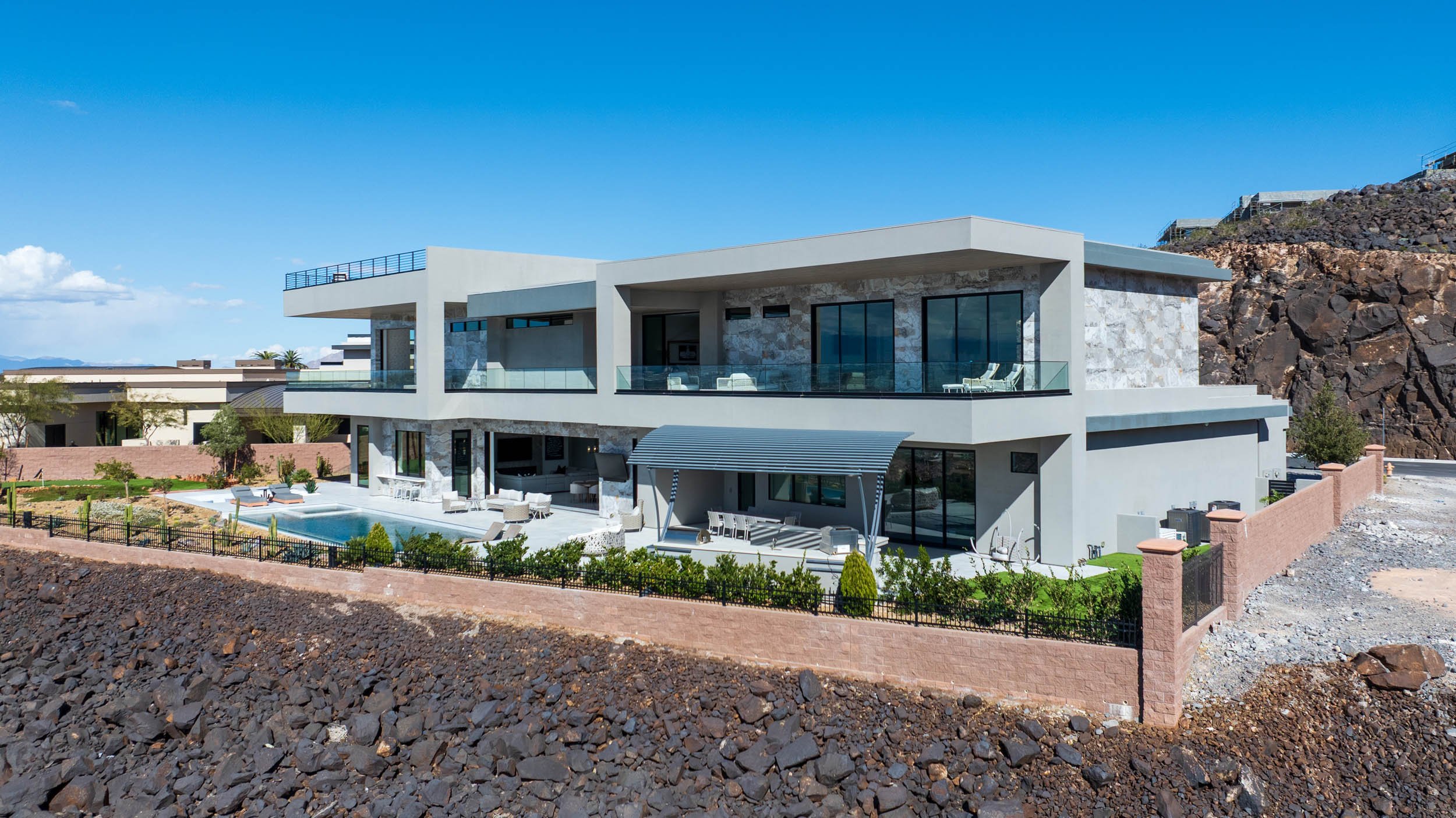 Modern multi-story house with large glass windows and a flat roof, located on a rocky hillside. The house has a spacious outdoor patio with seating, a swimming pool, and a landscaped yard enclosed by a low brick wall. Clear blue sky in the background
