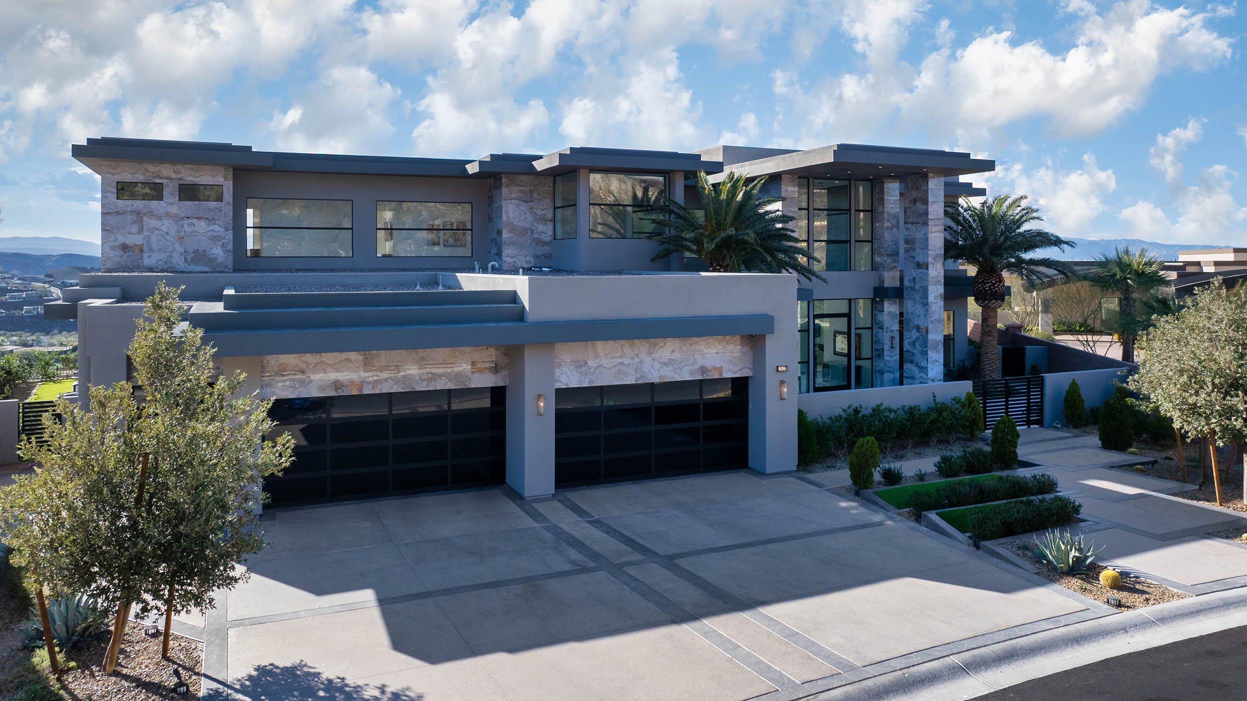 Modern house with a flat roof, large windows, and stone and stucco exterior, surrounded by desert landscaping and palm trees, under a partly cloudy sky.