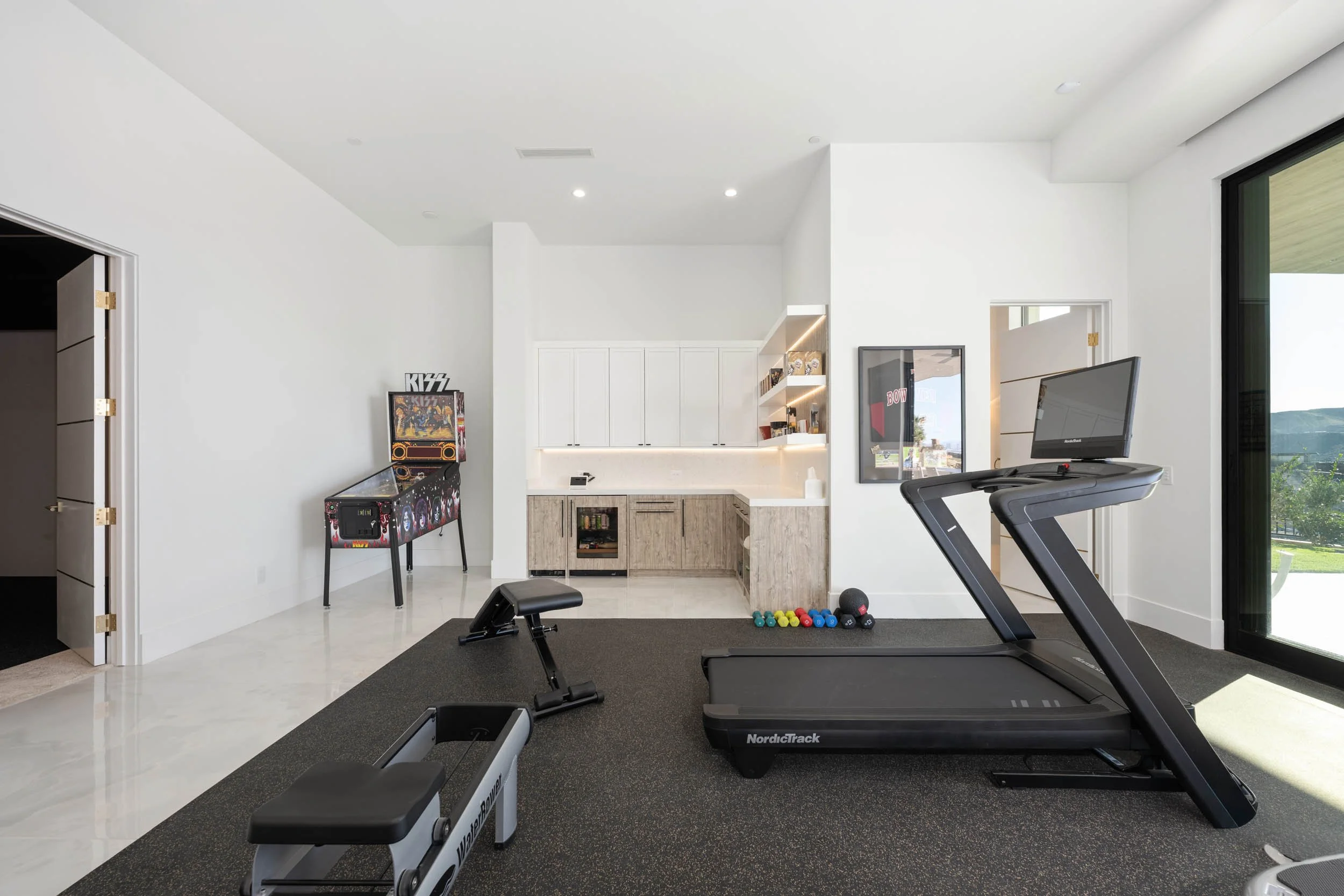 Home gym with treadmill, exercise bike, dumbbells, and snack bar area with a pinball machine and wall art.
