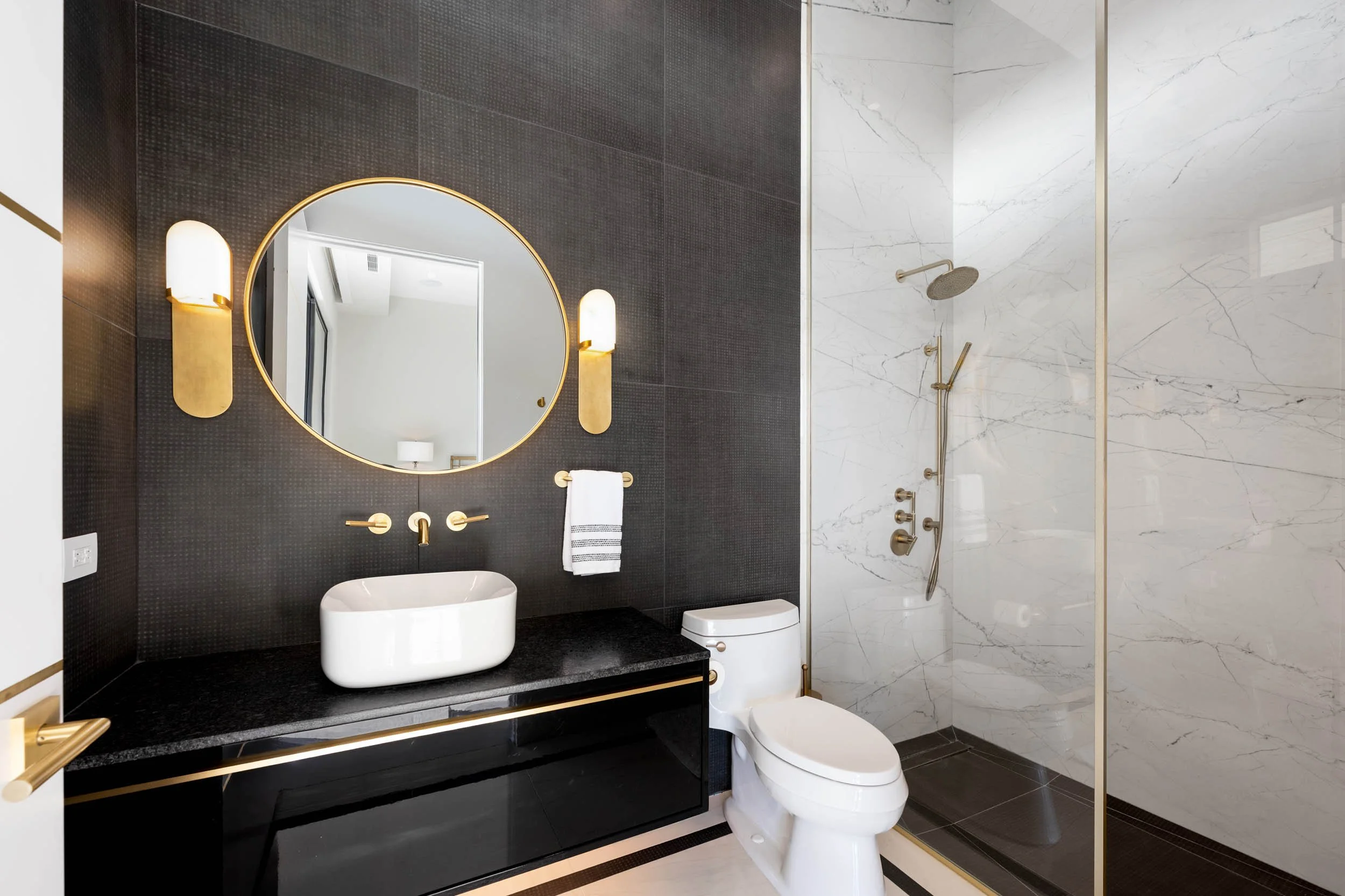 A modern bathroom featuring a black wall with a round mirror, gold accents, and wall lamps. There is a white vessel sink on a black countertop, a towel hanging on a gold bar, a white toilet, and a marble-tiled walk-in shower with gold fixtures.