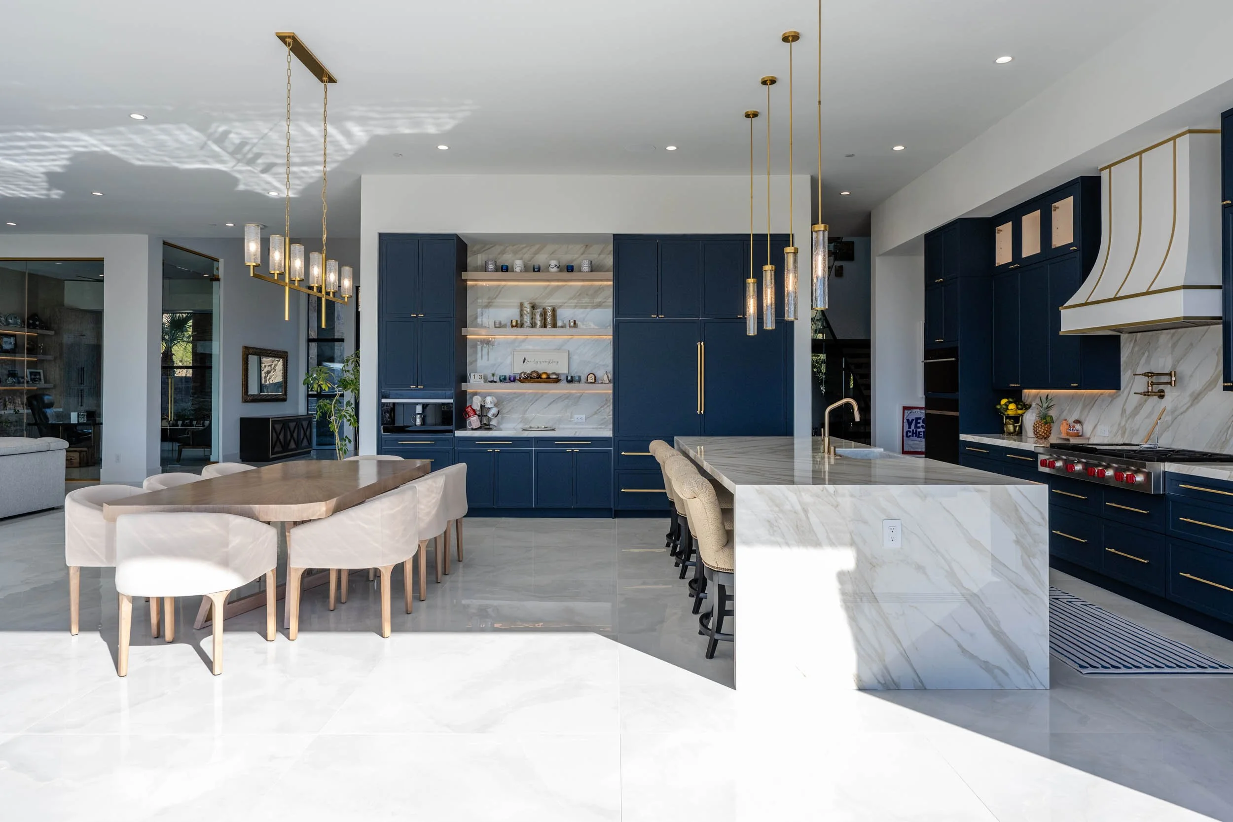 Modern kitchen with blue cabinets, white marble island and open shelving, pendant lighting, and a dining table with eight white chairs.