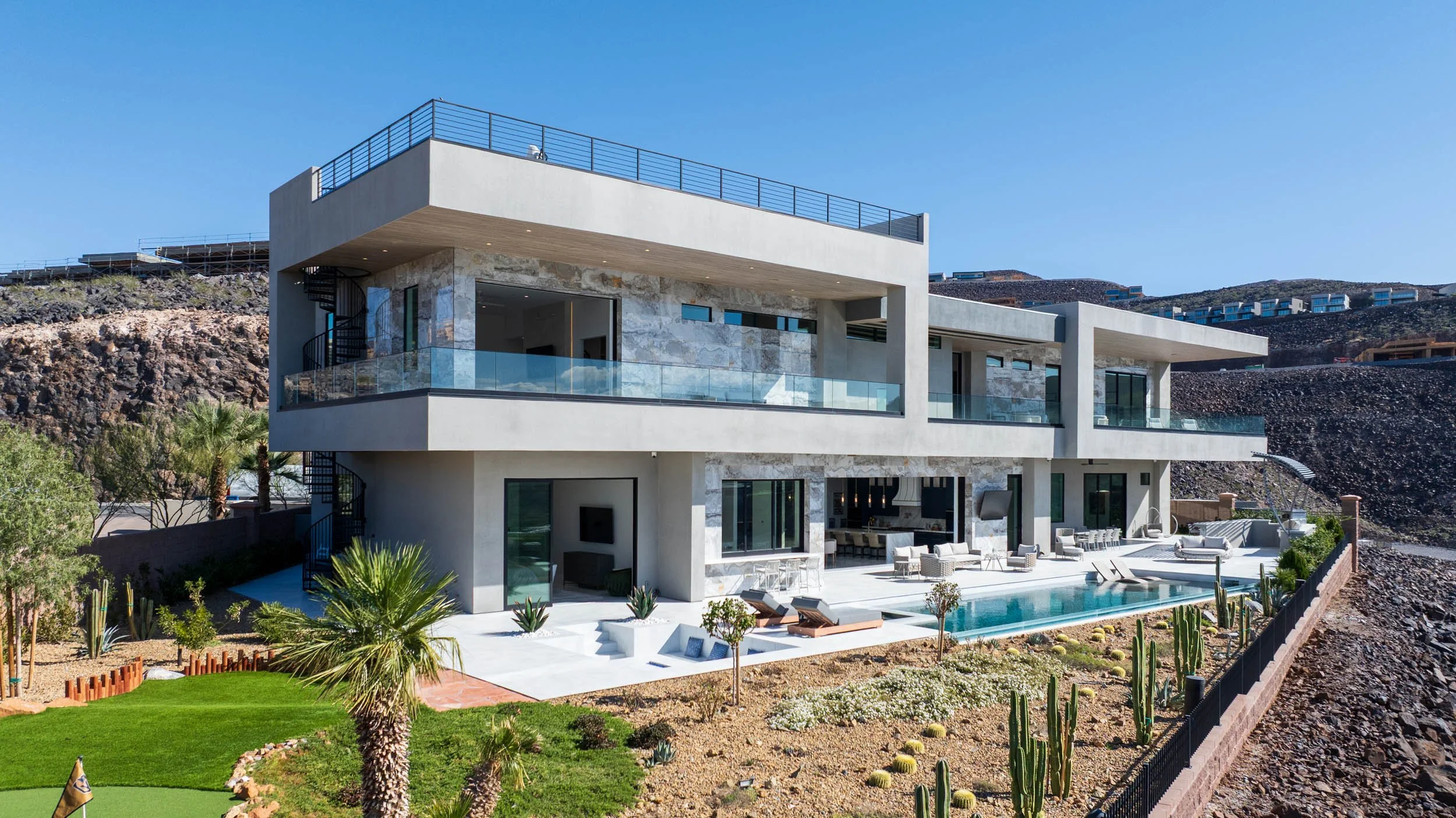 Modern multi-story house with large balconies, outdoor pool, patio furniture, desert landscaping, and hills in the background under a clear blue sky.