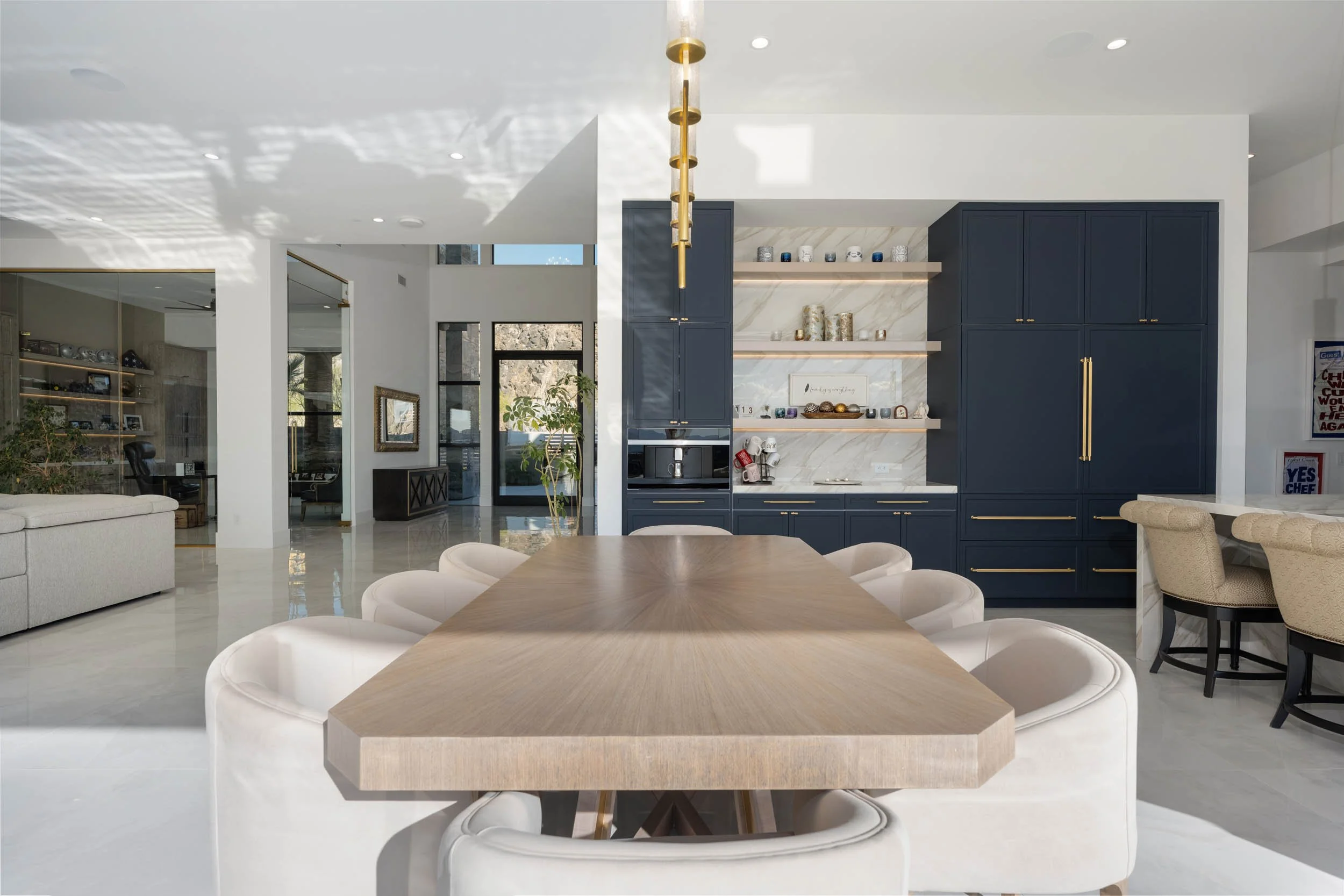 Modern open-concept kitchen and dining area with a large wooden table surrounded by white upholstered chairs, navy blue cabinetry, open shelves with decorative items, marble backsplash, and minimalist decor.