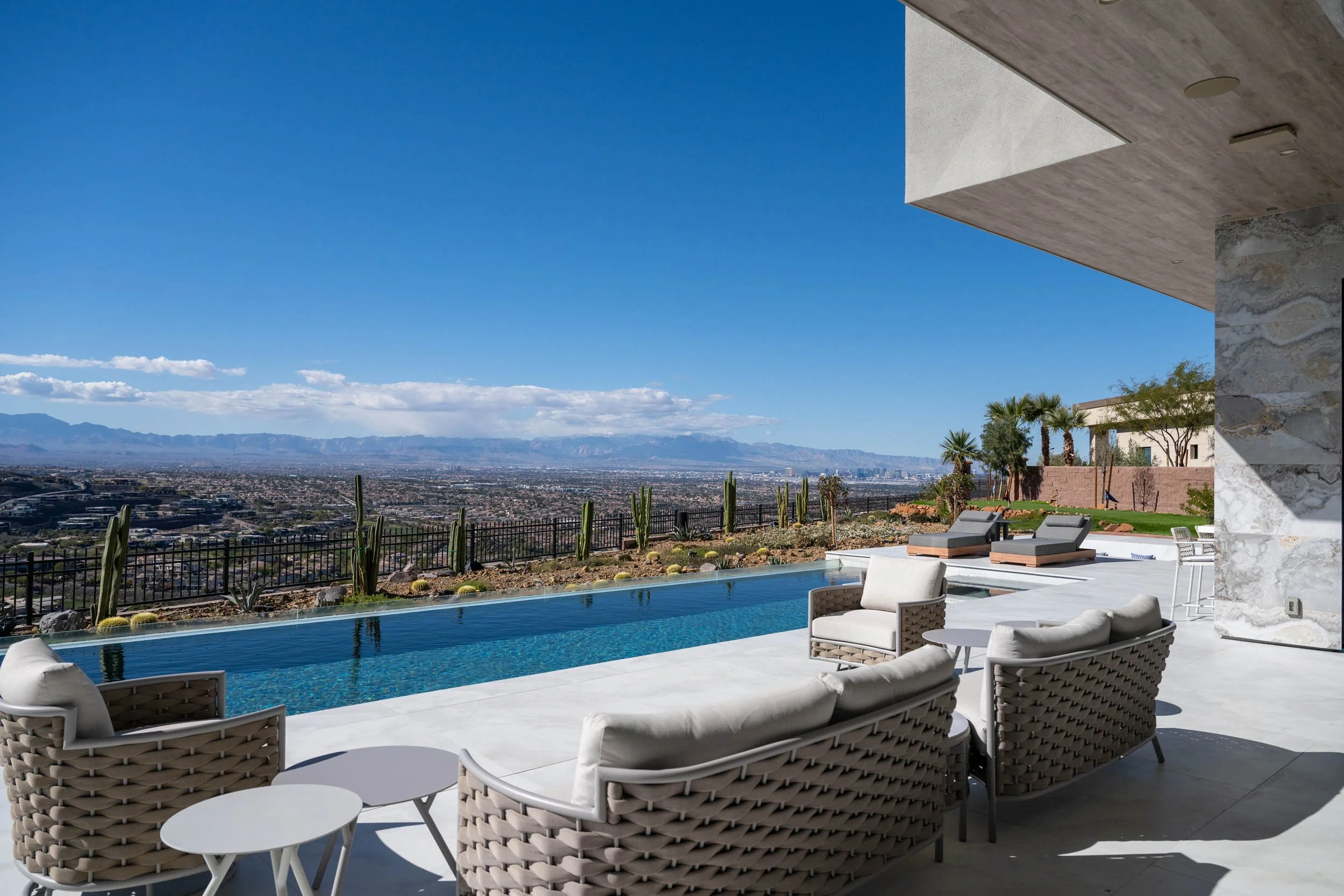 Modern outdoor patio with seating area, pool, desert landscape, and city view in the distance under a clear blue sky.