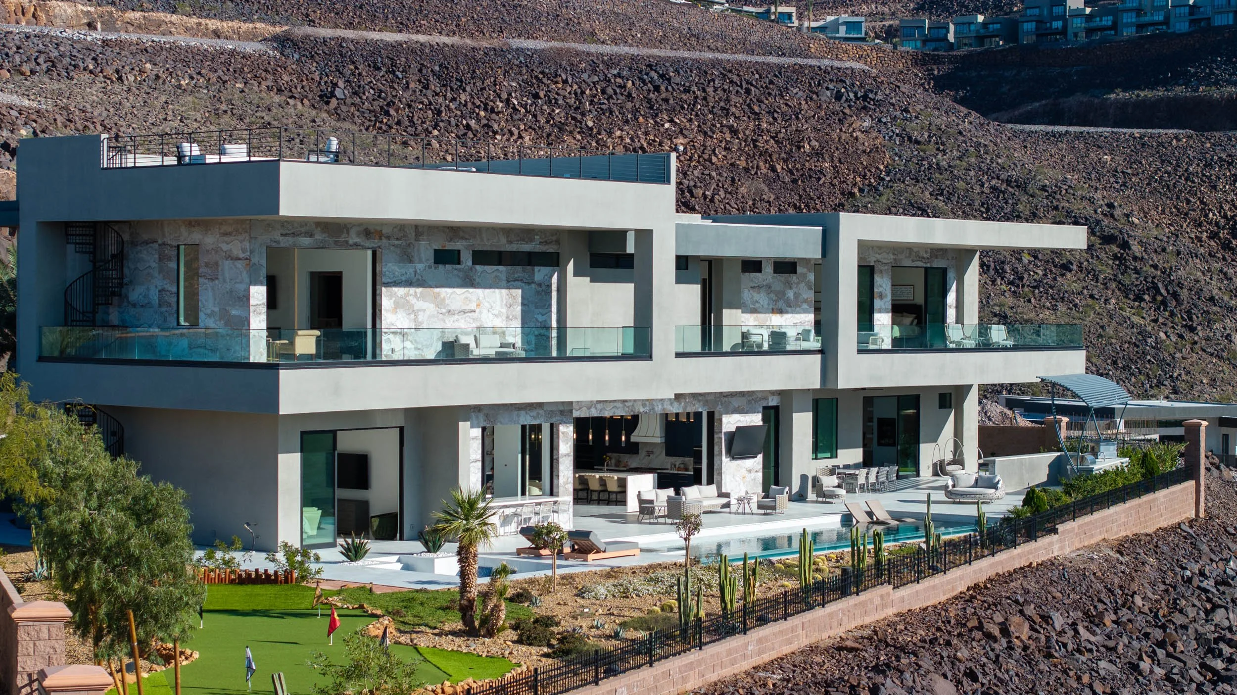 Modern multi-story house with outdoor lounge and pool, situated on a hillside with desert landscaping.