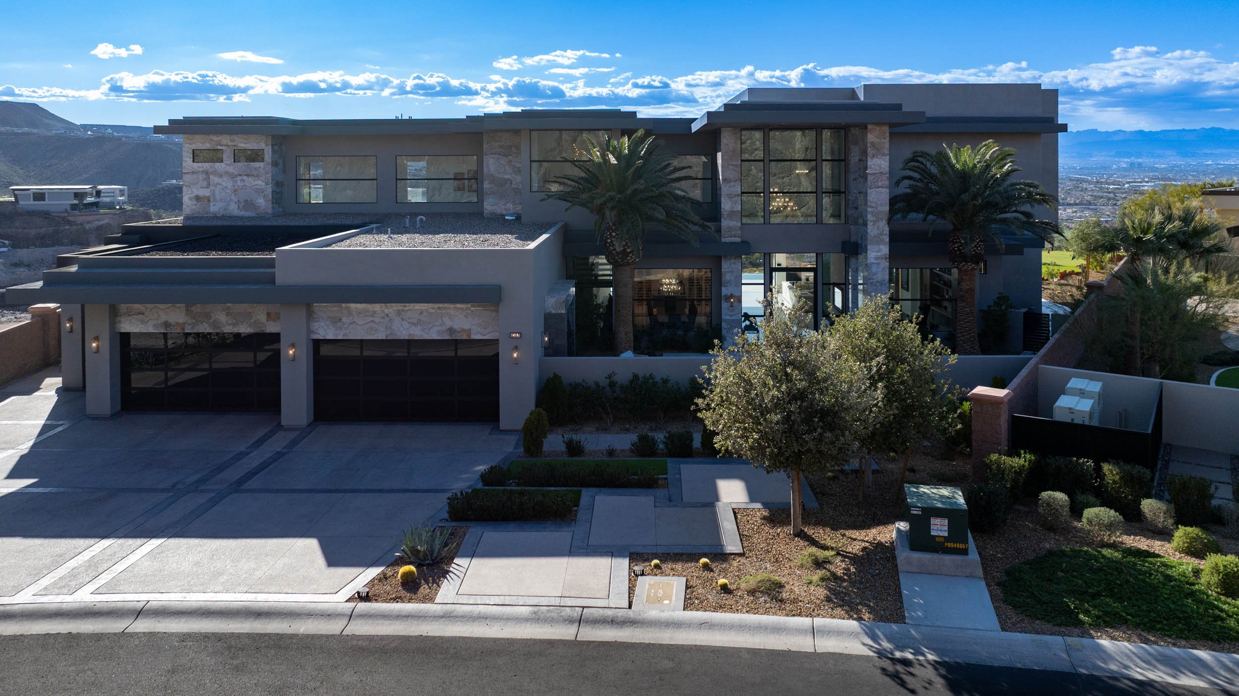 Modern two-story house with large glass windows, palm trees, and a desert landscape, situated in a hilly area under a partly cloudy sky.