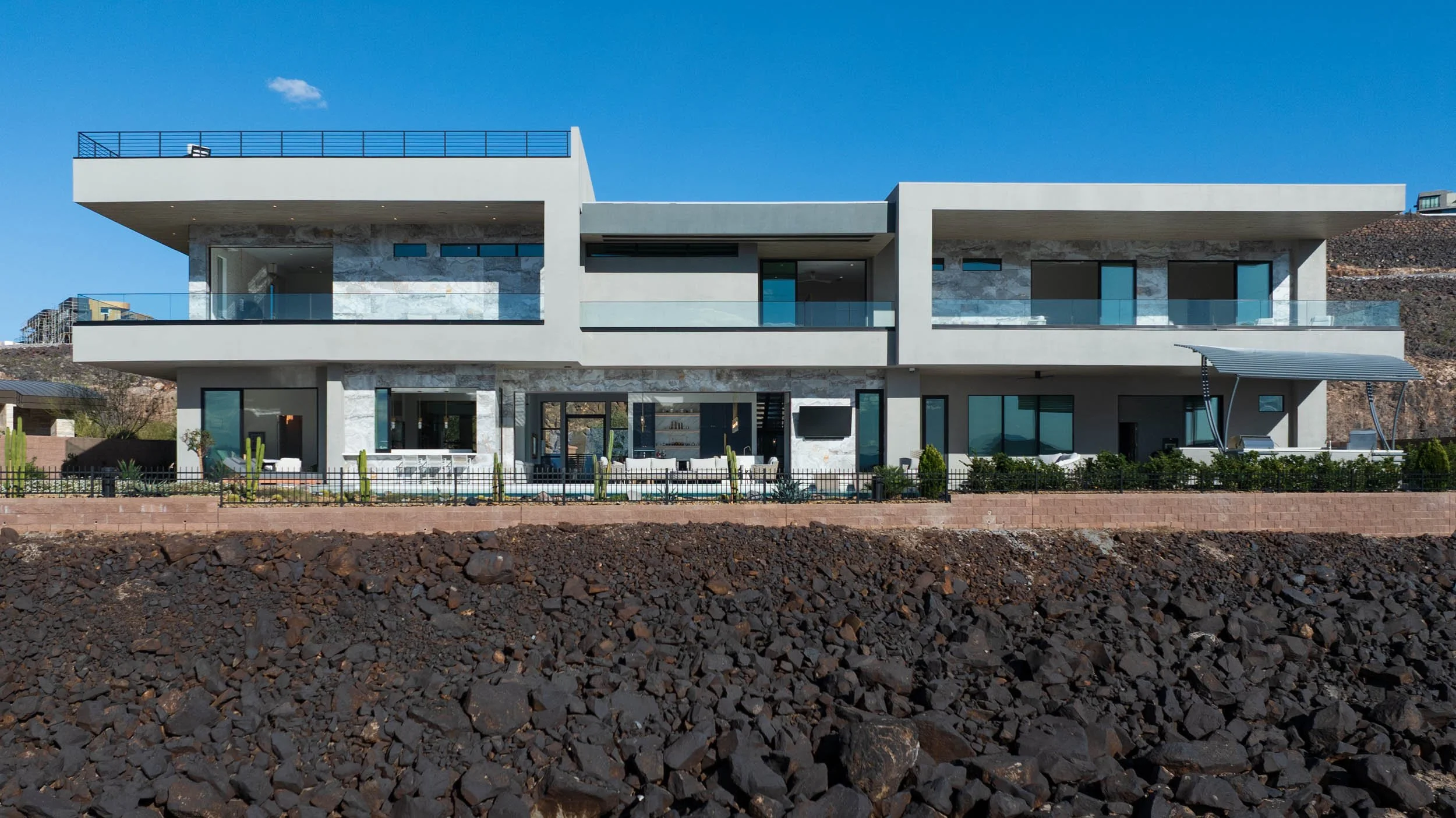 Modern multi-story house with large windows, outdoor balconies, and outdoor seating, set against a rocky landscape and clear blue sky.