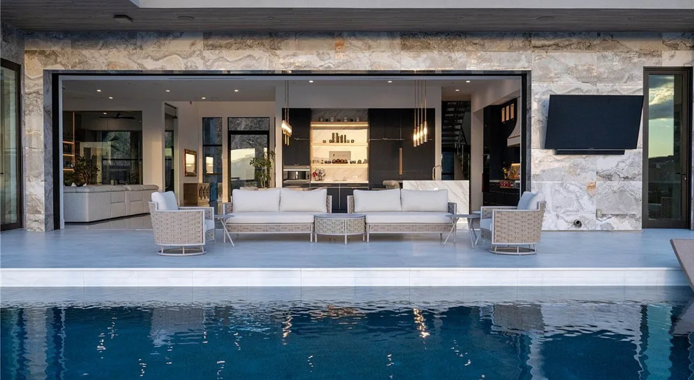Modern outdoor patio with a swimming pool, white furniture, and open view into a stylish kitchen and living area.