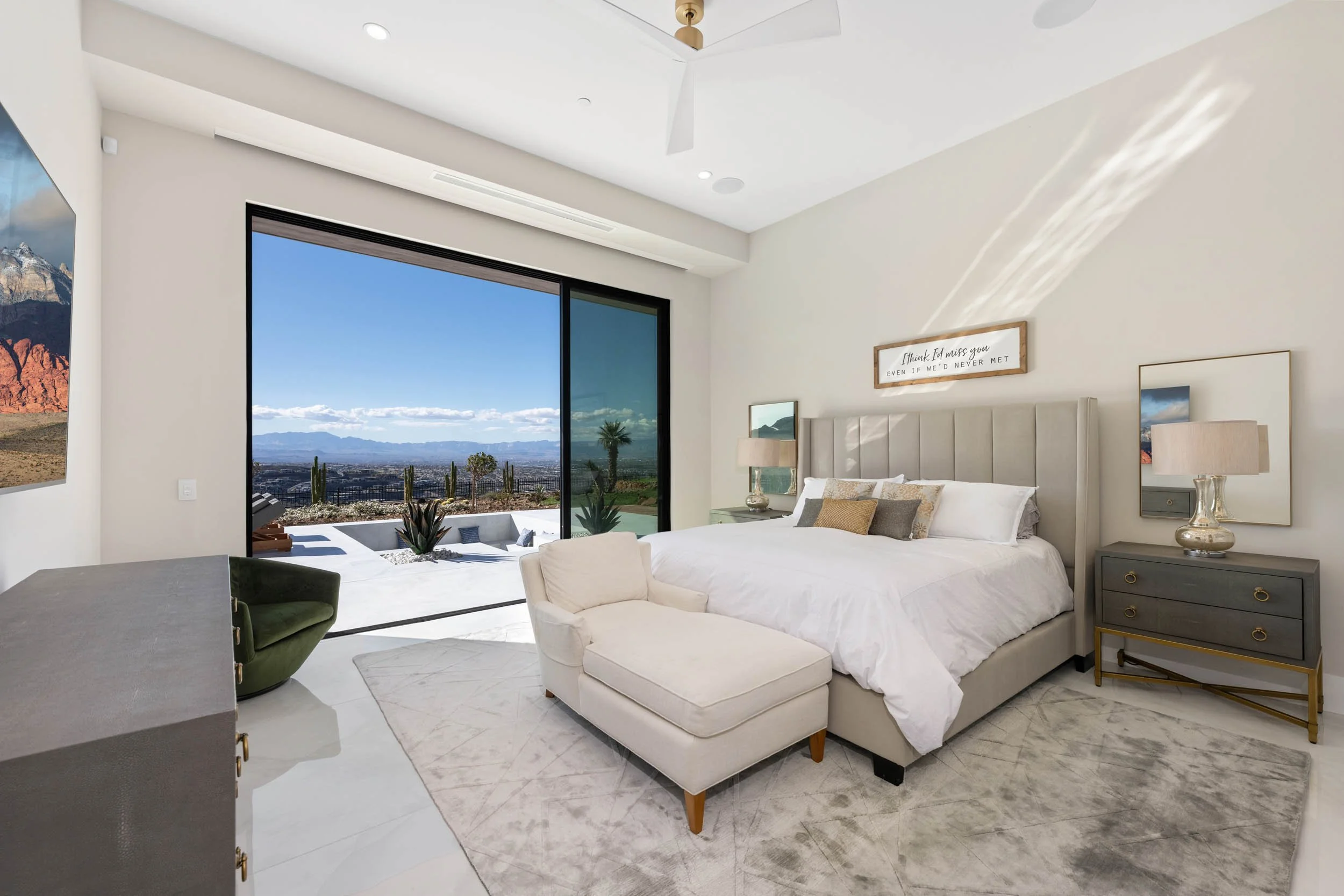Modern bedroom with large sliding glass door showing desert landscape and mountains outside, a beds and side tables with lamps inside, and sunlight casting shadow on the wall.