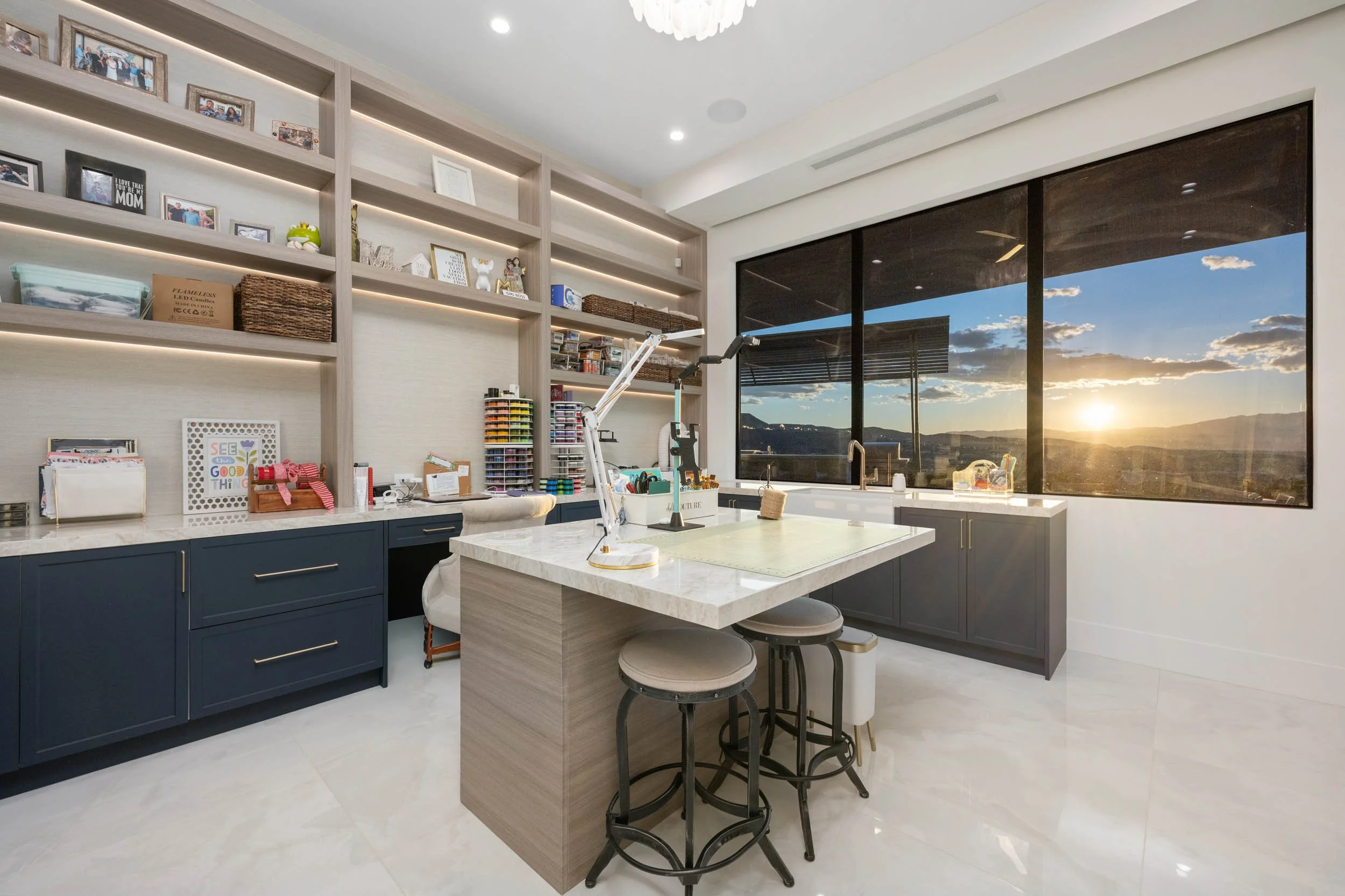 Modern craft or workroom with large window showing sunset over mountains, shelves with framed photos, art supplies, and a workspace with a marble-topped island.