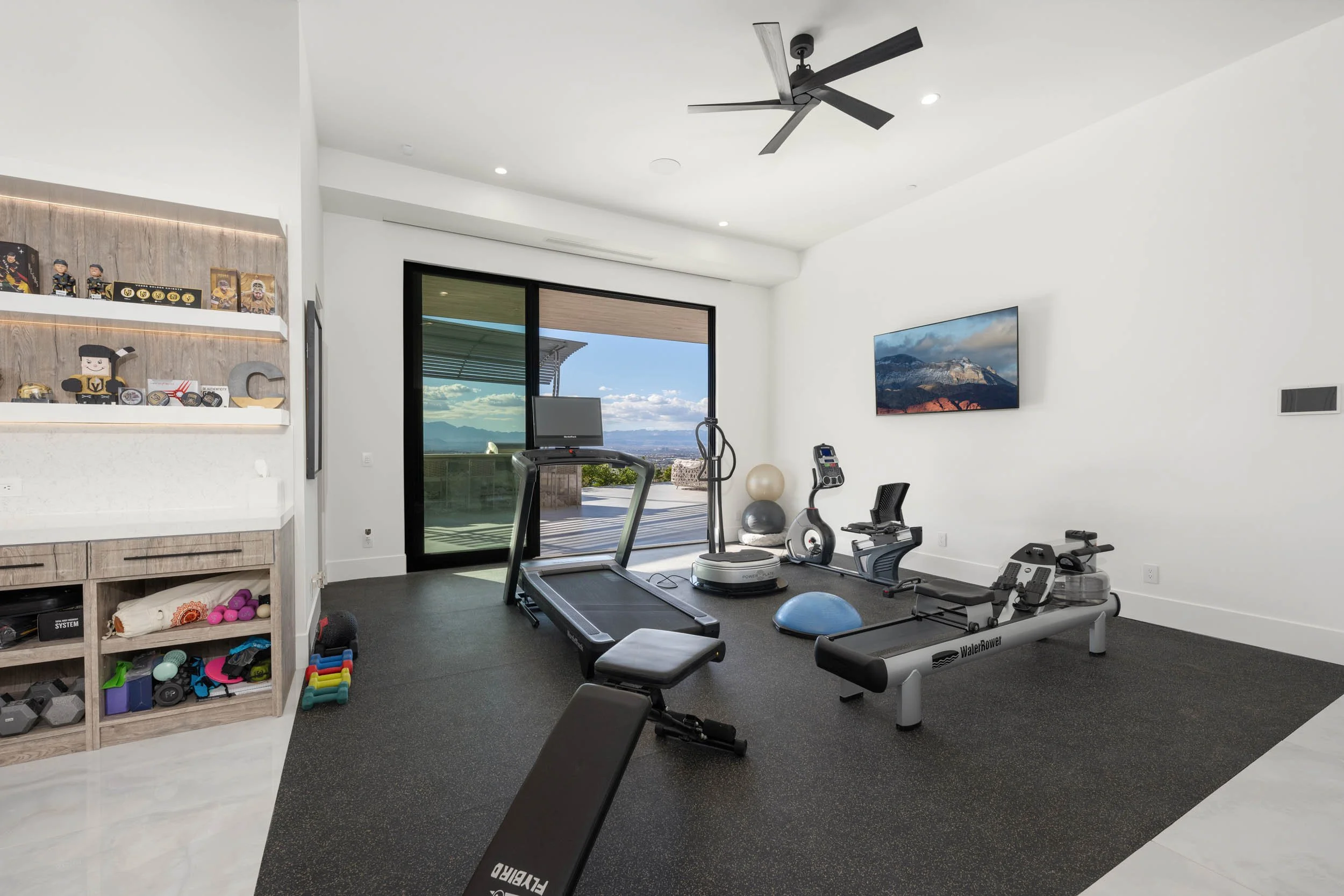 Home gym with treadmill, stationary bike, rowing machine, stability ball, exercise bike, and fitness accessories, with a large sliding door overlooking an outdoor balcony and scenic mountain view.