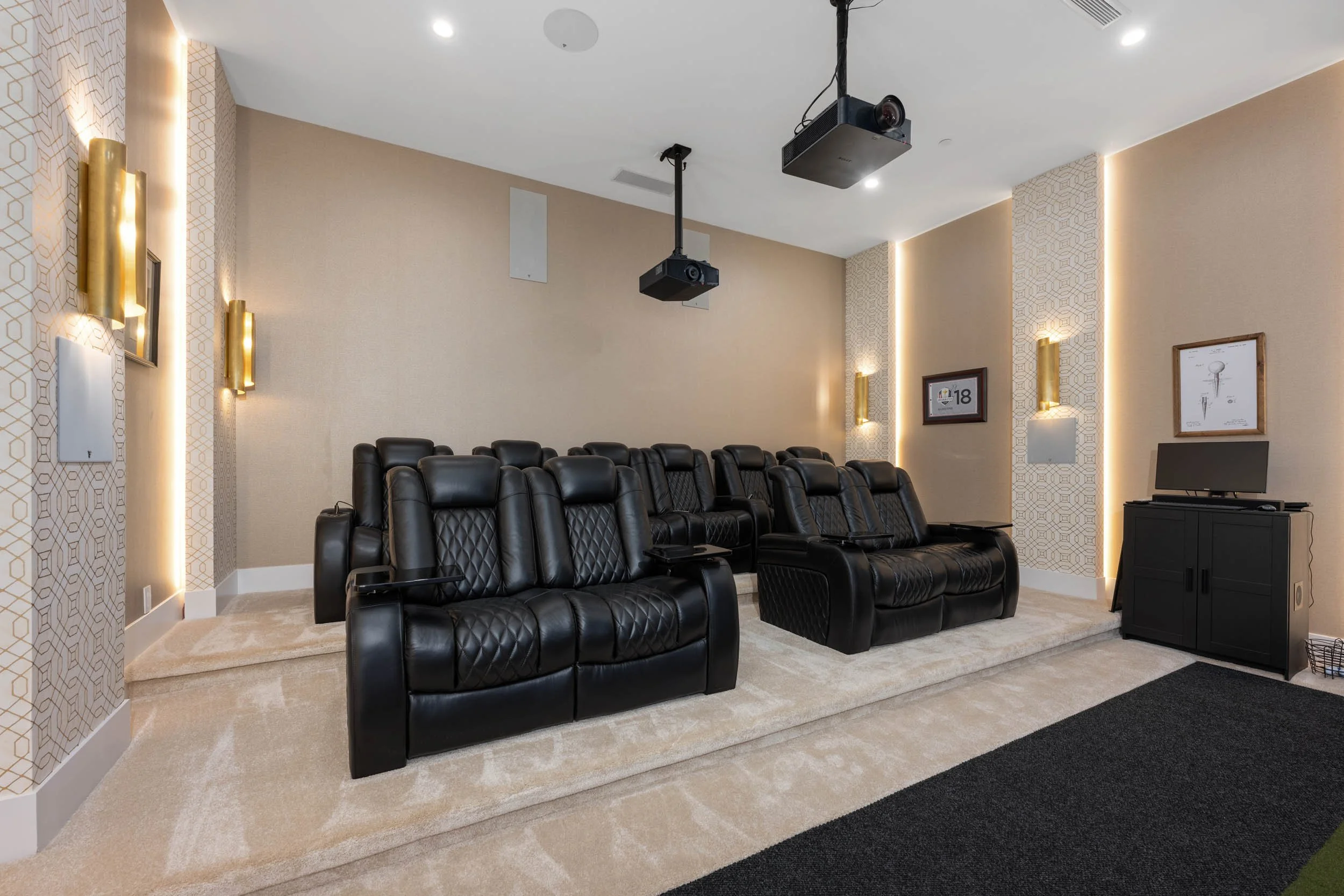 Home theater room with beige walls, black leather reclining chairs, wall-mounted gold and white light fixtures, framed pictures, a ceiling-mounted projector, a black cabinet with a monitor, and a beige carpeted floor with a black rug.