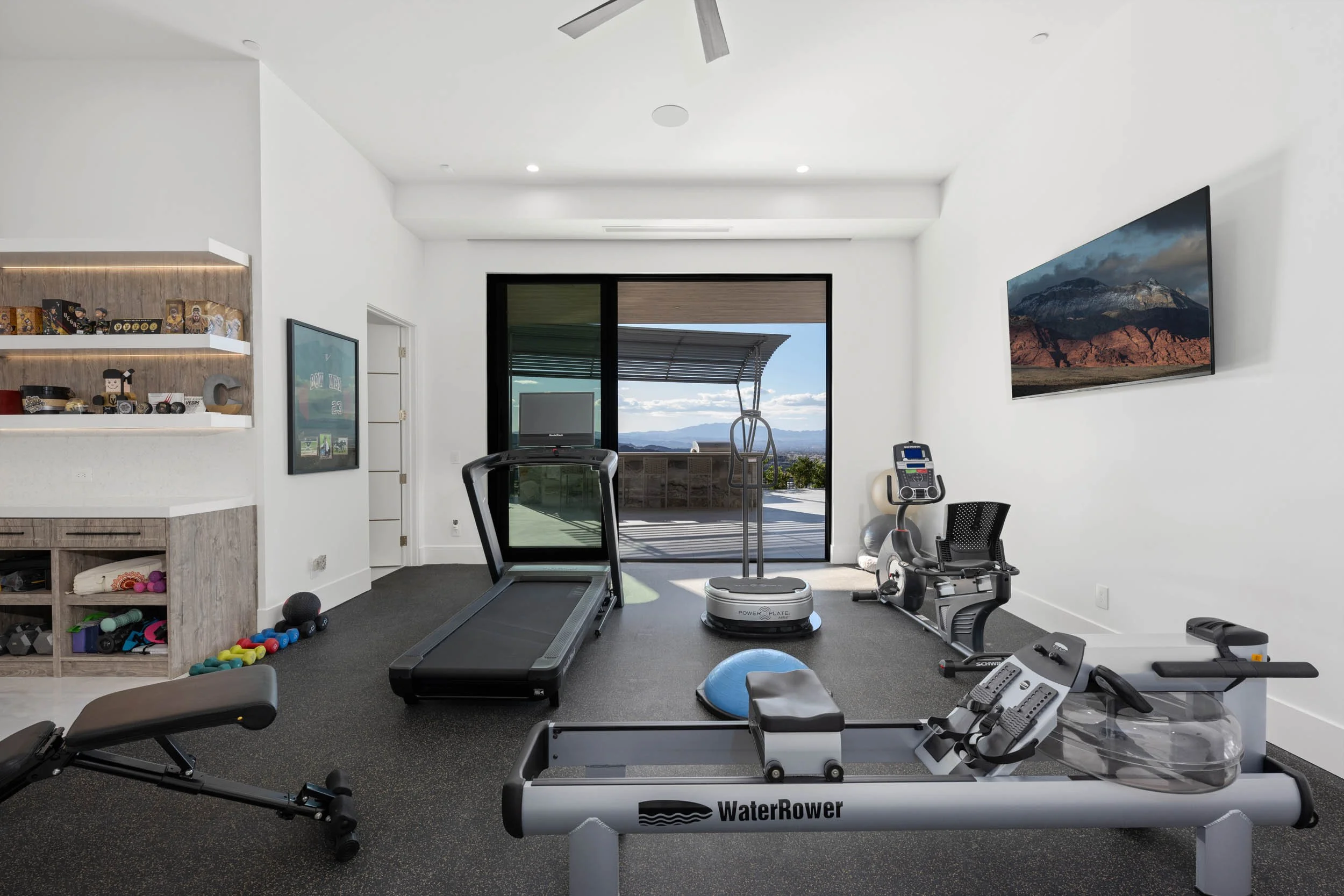 A home gym with treadmill, stationary bike, WaterRower rowing machine, exercise ball, and various weights, with a large sliding glass door opening to an outdoor patio with mountain view