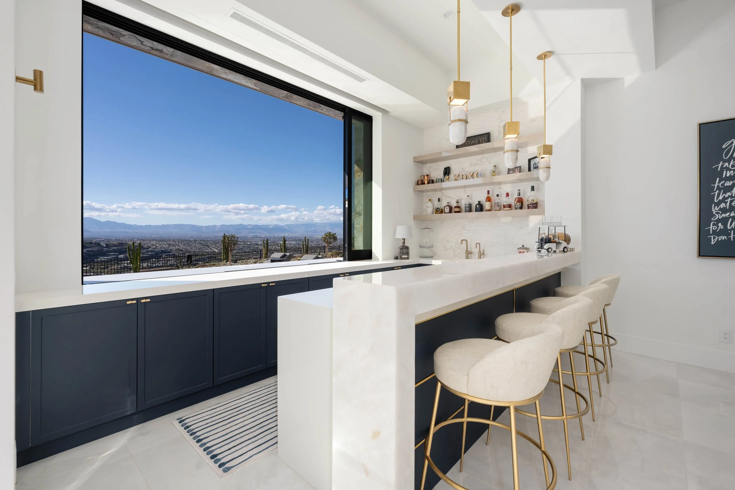 Modern kitchen with white marble island, blue cabinets, gold accents, barstools, open shelves with liquor bottles, large window with mountain view.