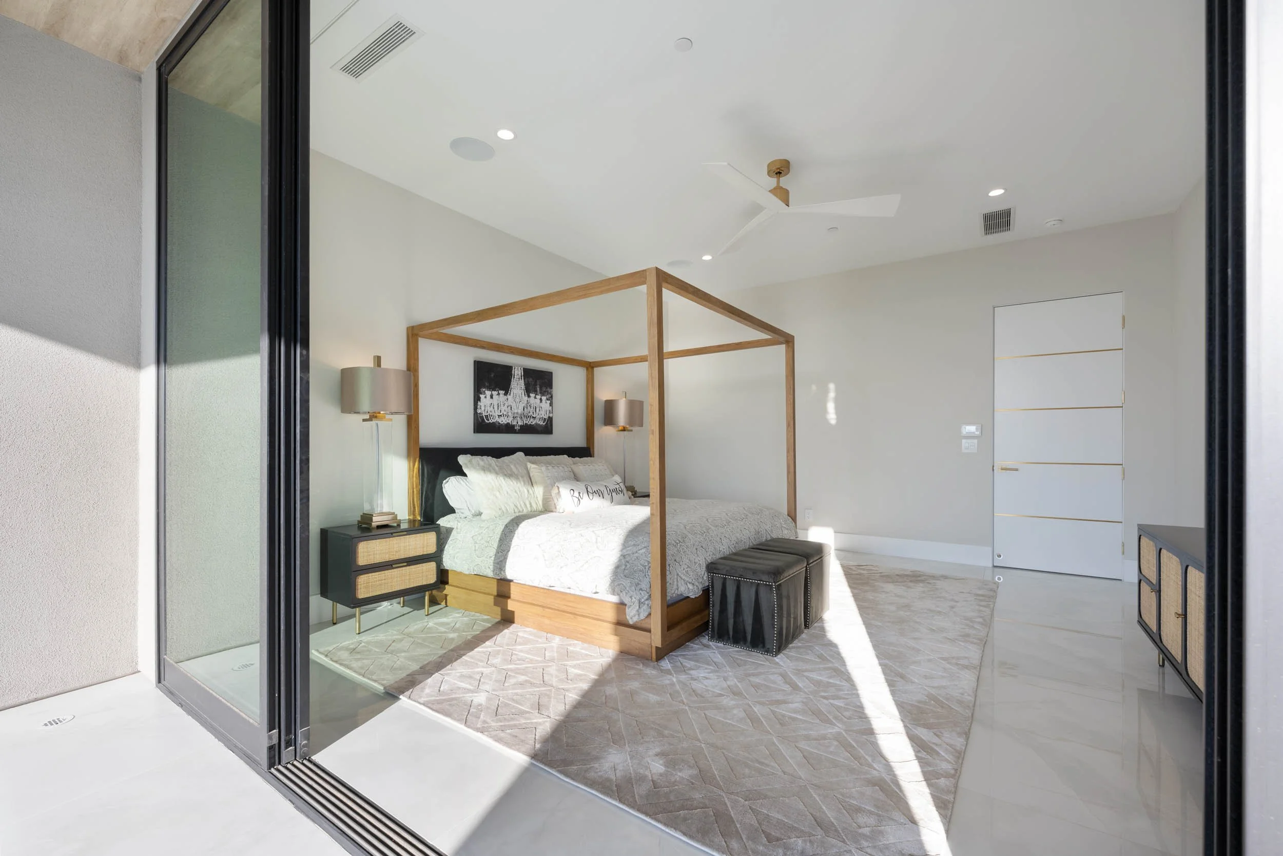 Modern bedroom with a four-poster bed, black and gold nightstands, and a large beige area rug, seen through a sliding glass door.