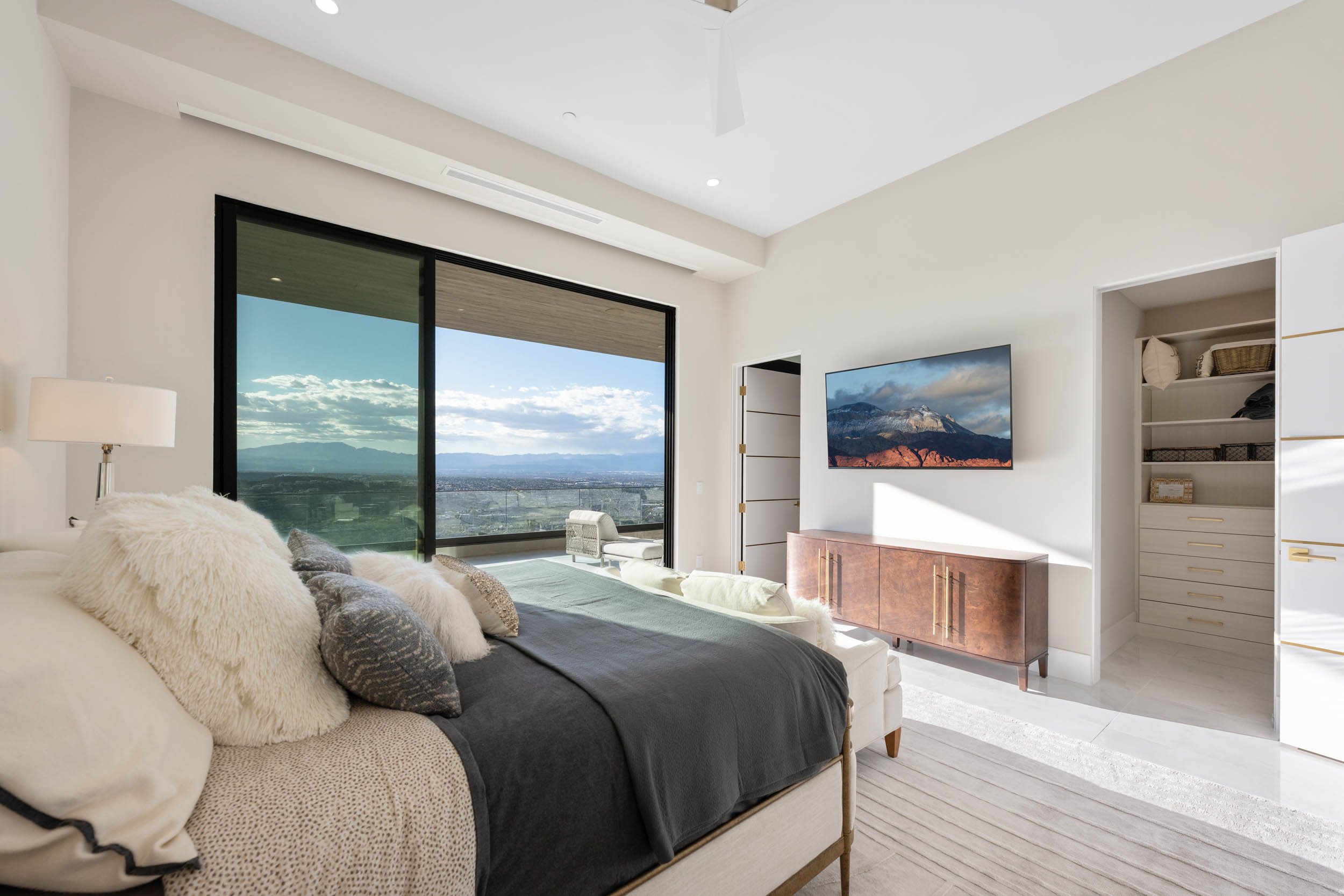 Modern bedroom with large window showing cityscape and mountains, white walls, TV on wall, white closet, and beige furniture.