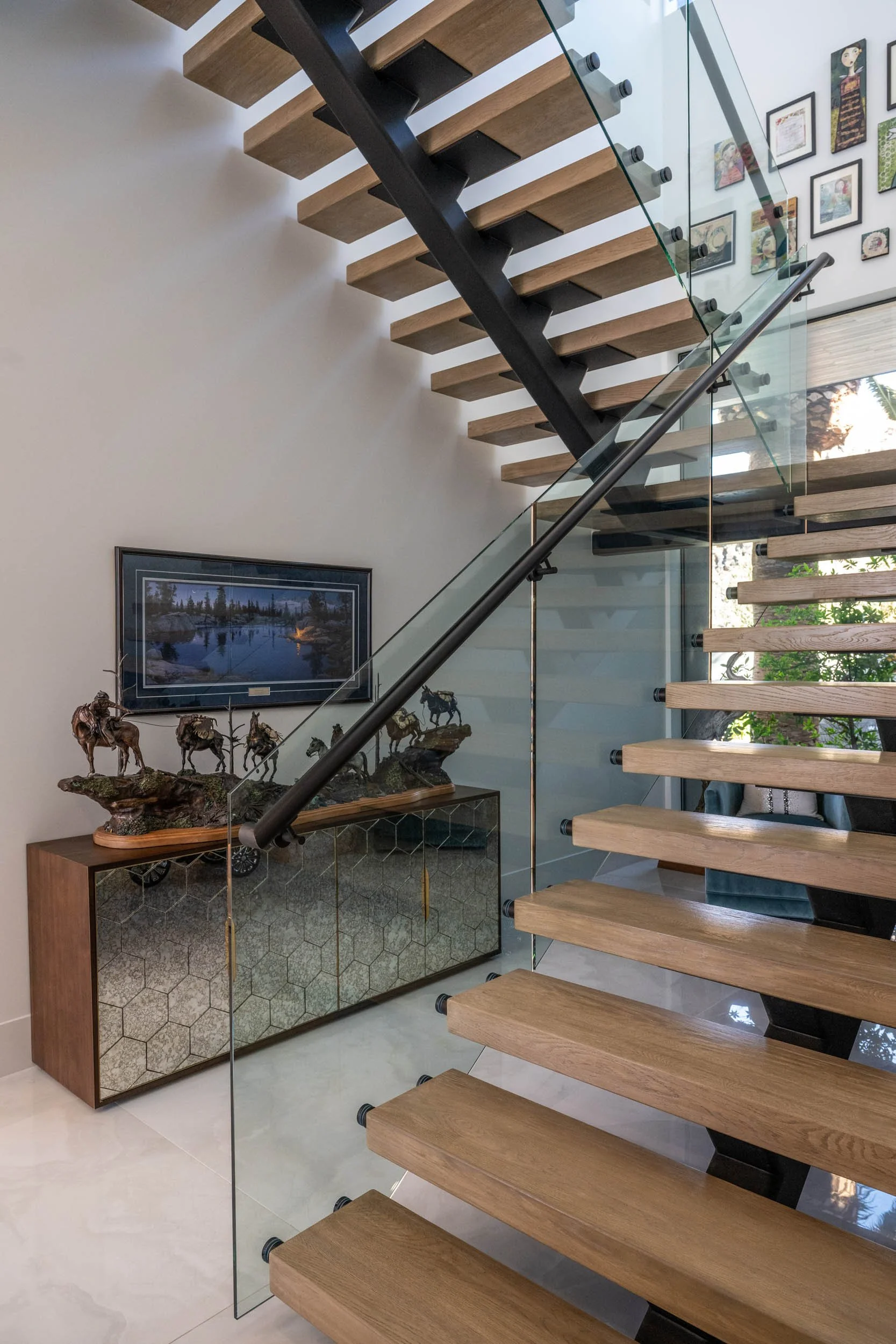Wooden staircase with glass railing in a modern interior, decorated with sculptures of horses on a wooden cabinet, and framed artwork on the wall.