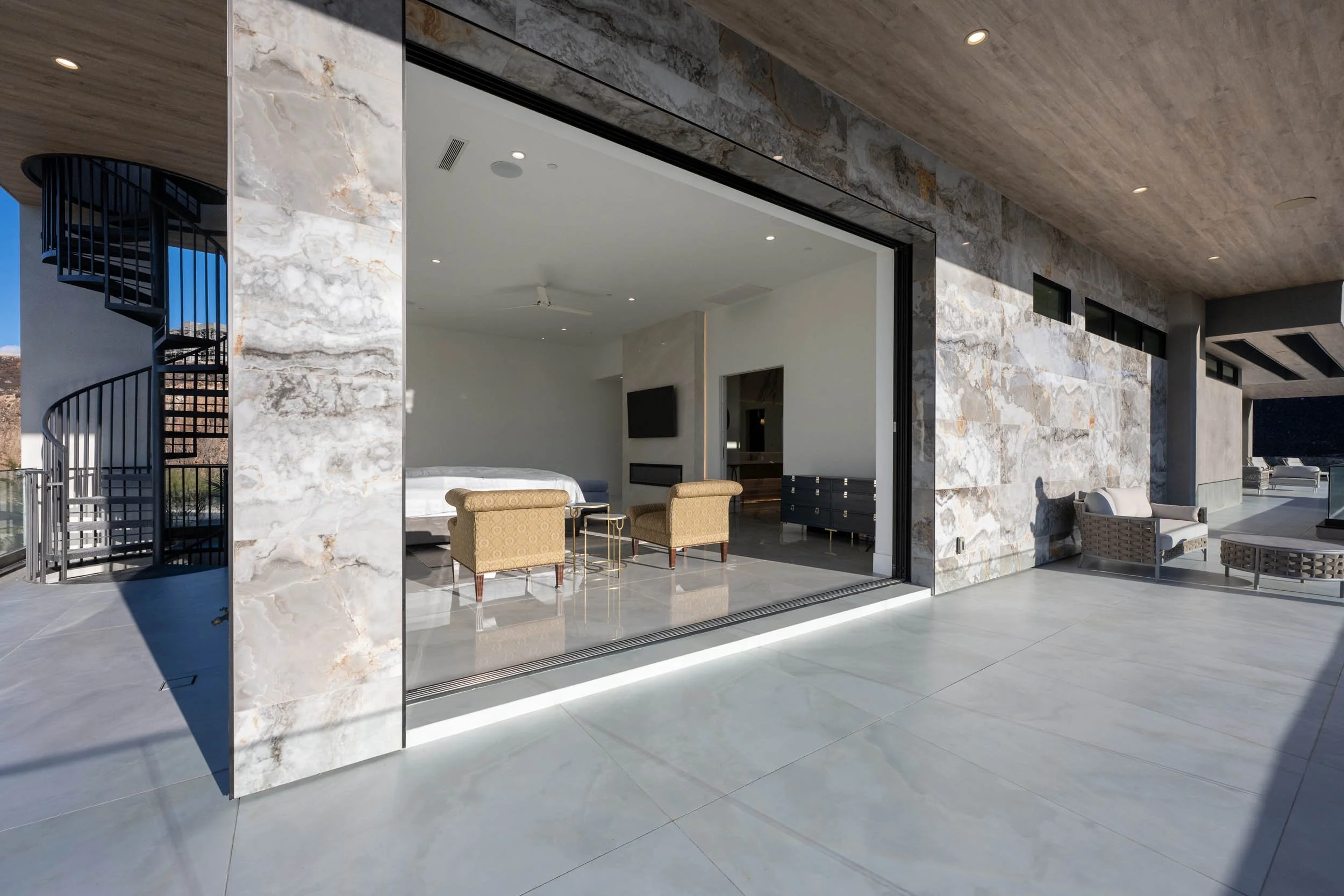Modern open-concept bedroom with sliding glass doors opening onto a spacious balcony, featuring contemporary furniture and natural stone exterior walls.
