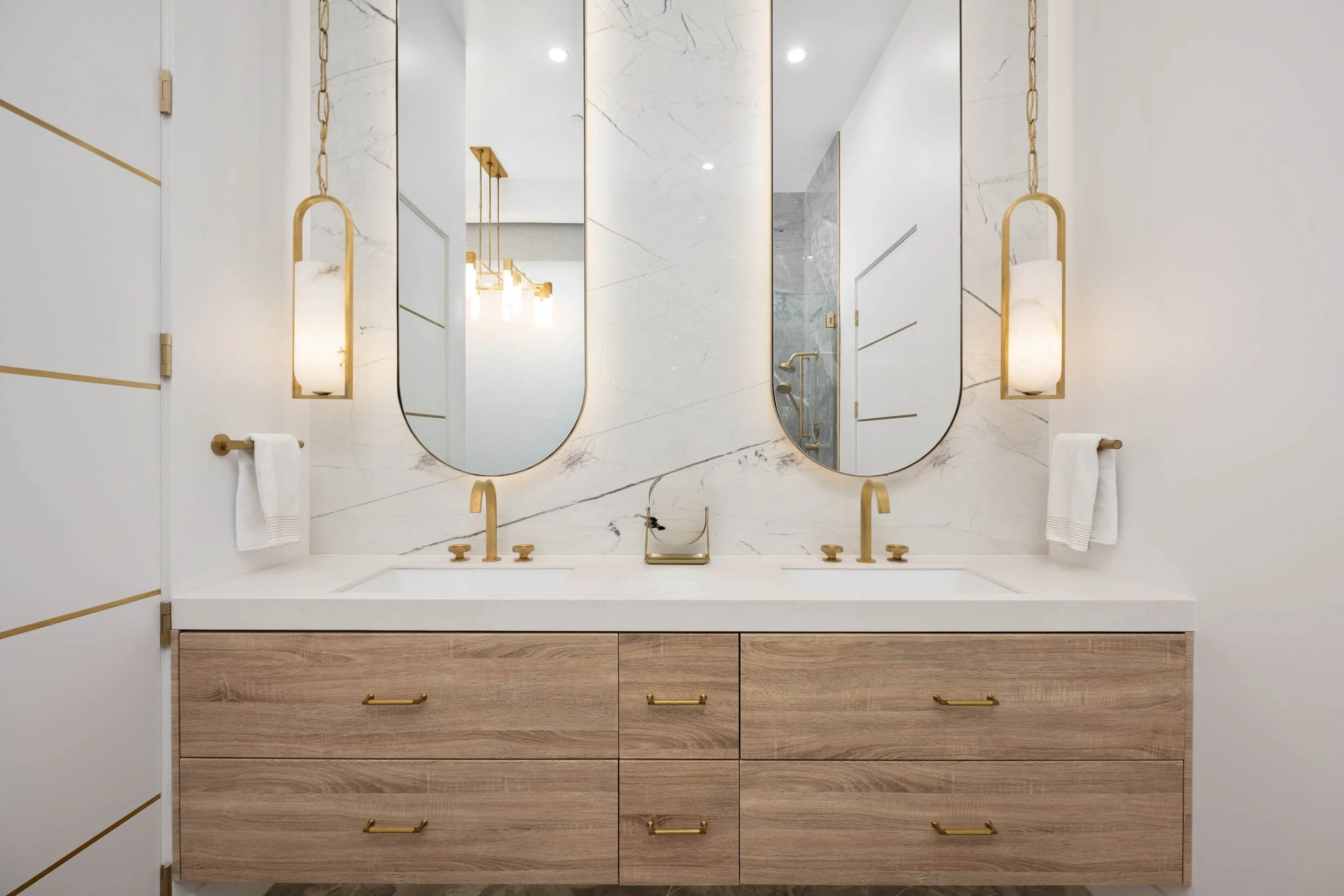Modern bathroom vanity with two oval mirrors, gold fixtures, and a marble backsplash, featuring a wooden cabinet with six drawers and white towels.