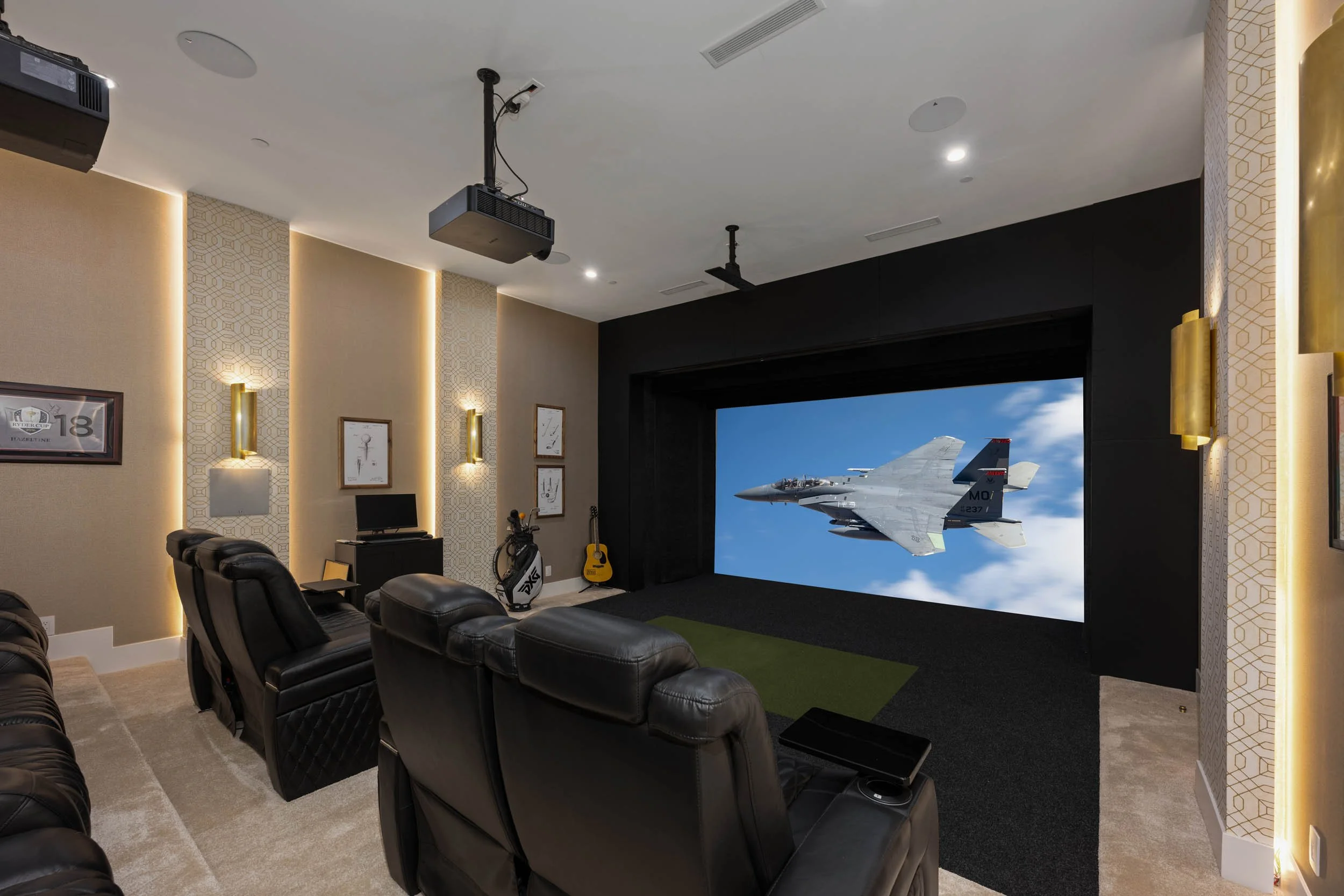 Home theater room with large screen showing a jet airplane in flight, black leather recliners, a golf bag, a guitar, and framed artwork on the wall.