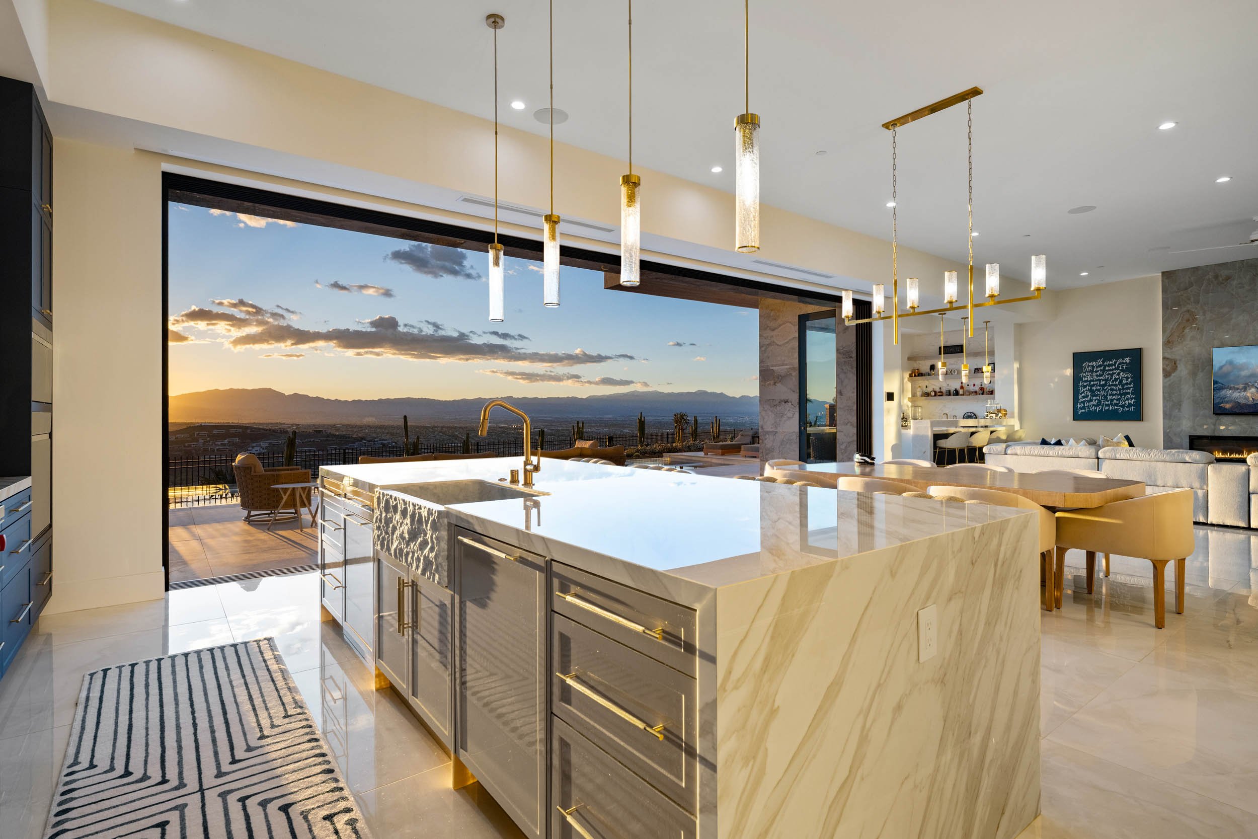 Modern open-concept kitchen and living room with large windows overlooking a sunset landscape, featuring a marble kitchen island with gold accents and contemporary pendant lighting.