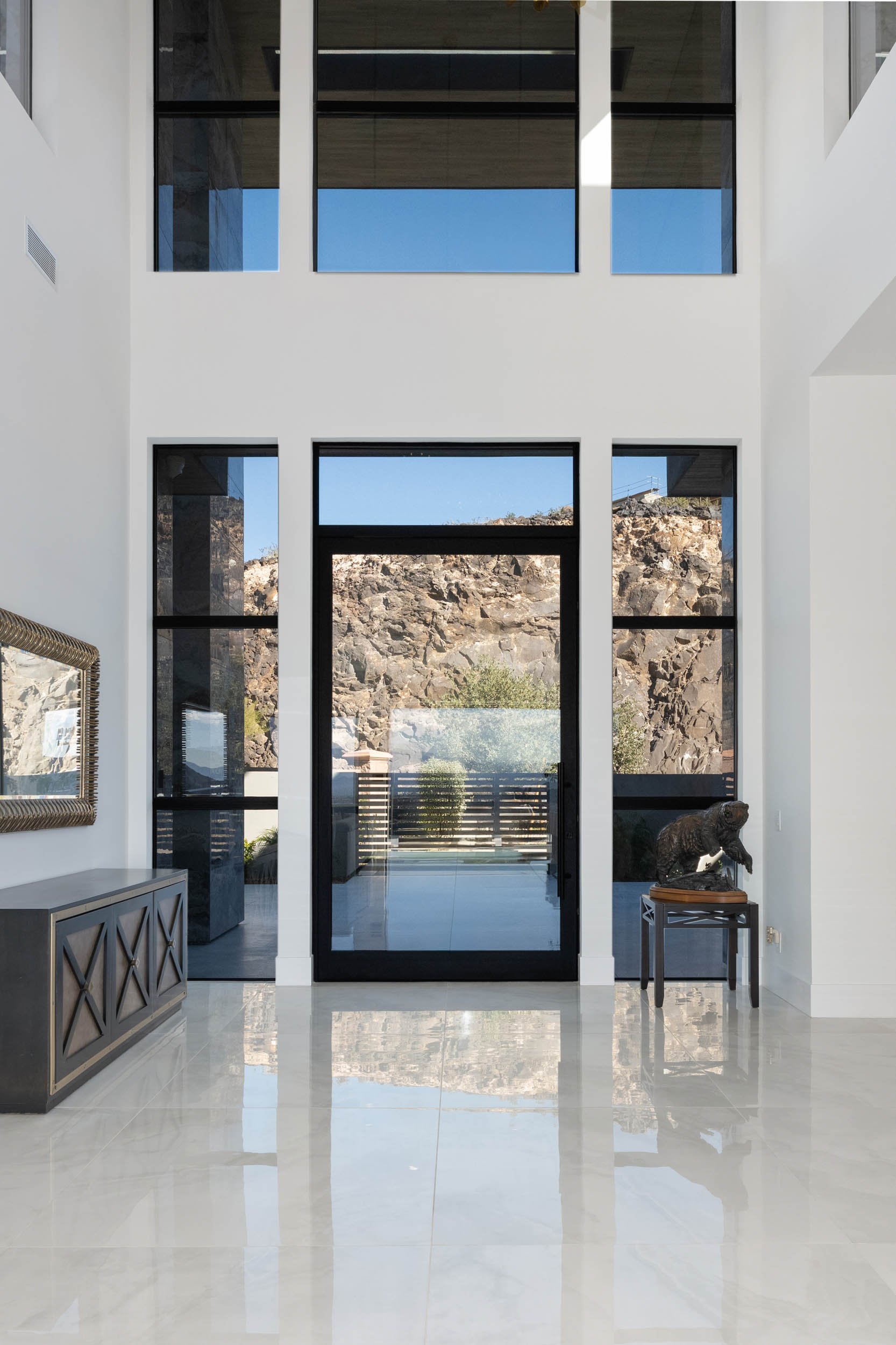 Modern interior with large glass windows and door, white walls, marble floor, mountain view outside, and a small sculpture of a bear on a table to the right.