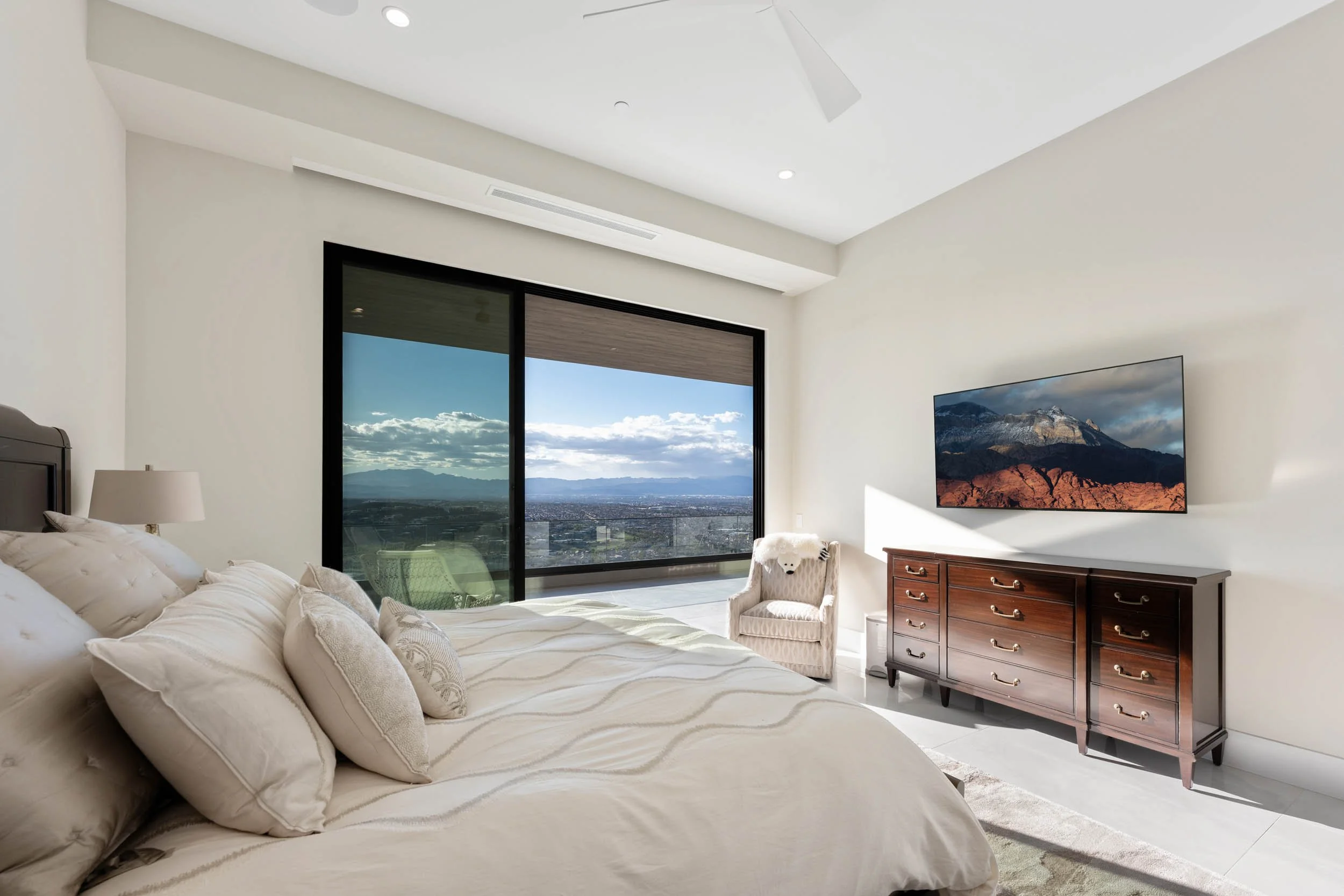 Modern bedroom with a large bed, a dresser, a mounted flat-screen TV, and a sliding glass door leading to a balcony with a view of mountains and a cloudy sky.